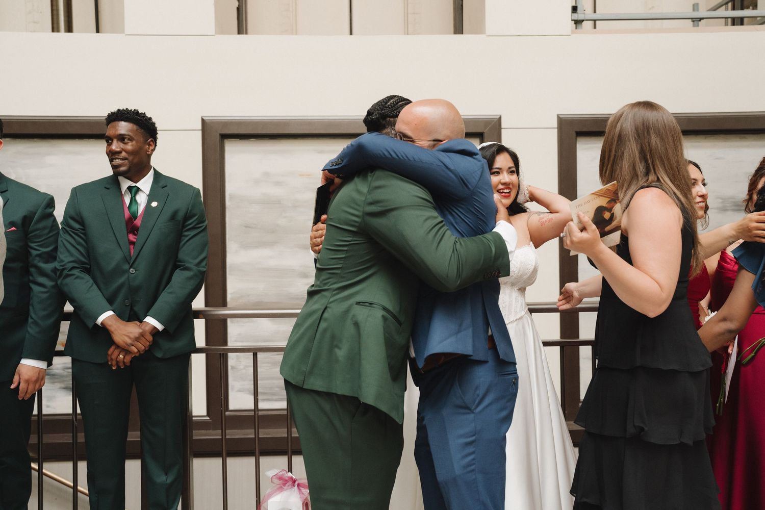 Two guests share a warm hug at a wedding celebration while others in formal attire look on and smile.