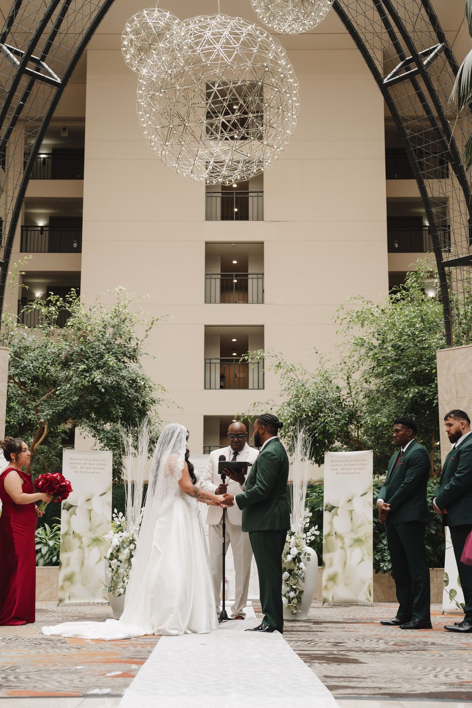 Bride and groom exchange vows beneath stunning spherical chandeliers in a grand hotel atrium wedding ceremony.