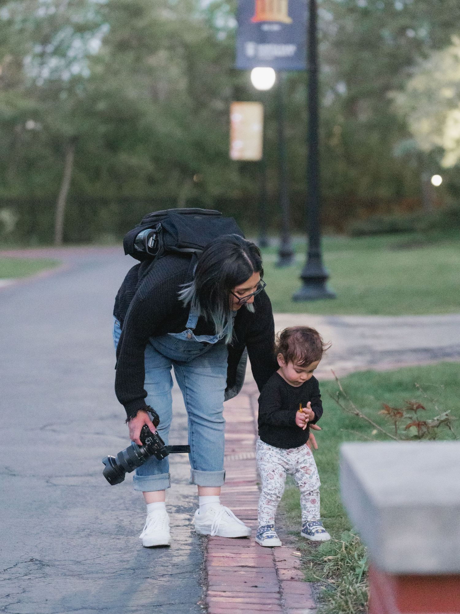 A photographer with a camera and backpack bends down to assist a toddler on a brick path in a park at dusk.