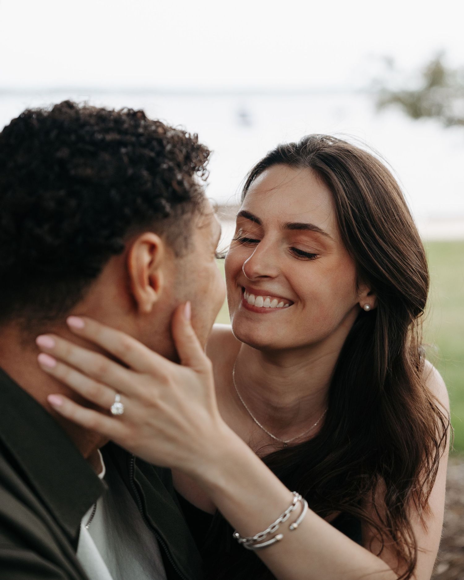 A smiling woman with a diamond engagement ring gently cups a man's face outdoors near water.