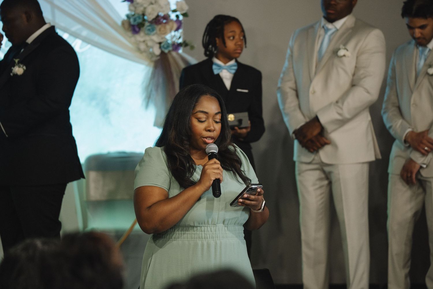 A woman in a mint dress reads from a phone into a microphone at a wedding ceremony.