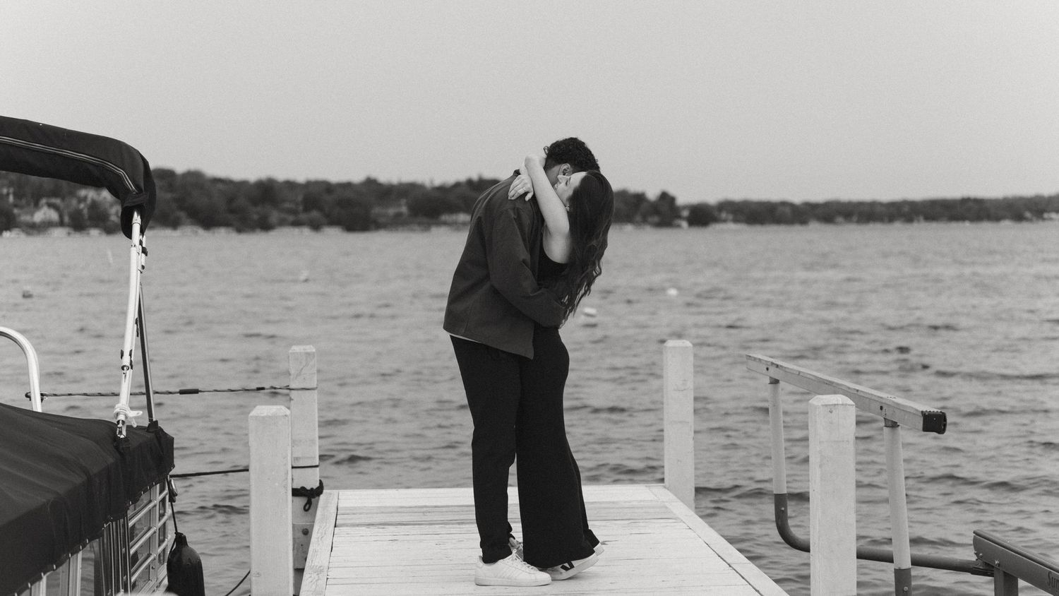 Couple embraces on dock by lake in black and white romantic engagement photo.