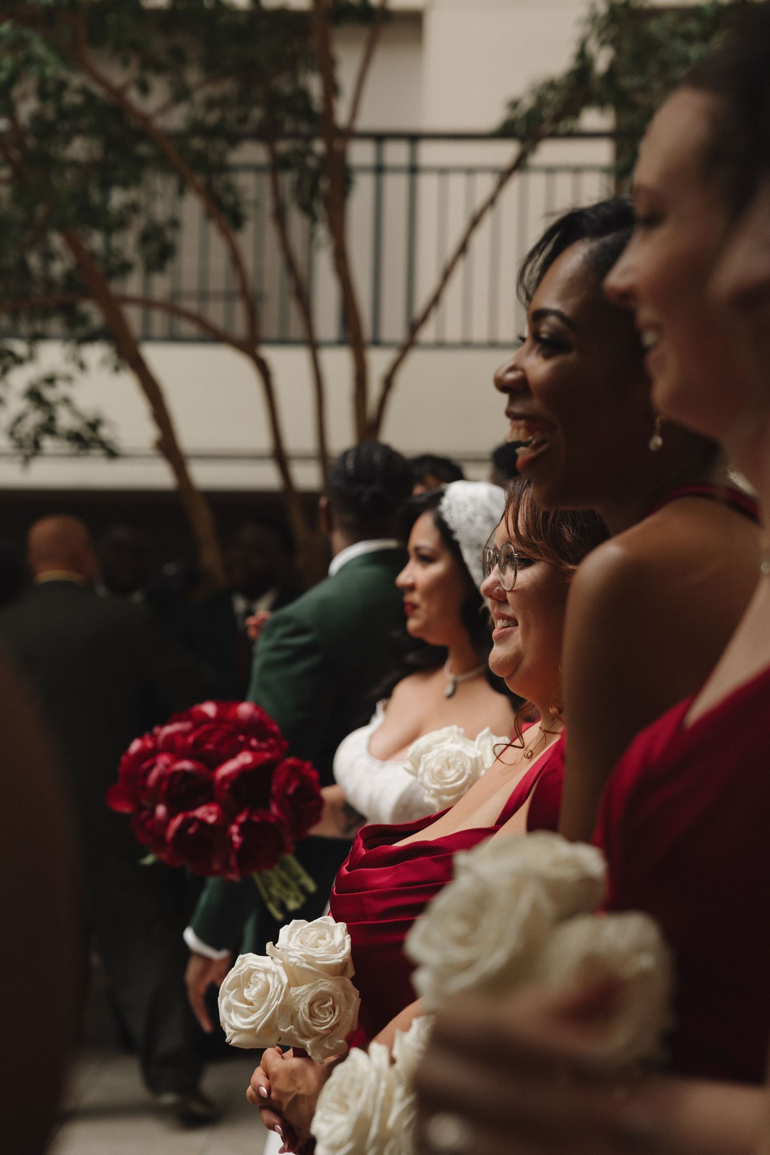 Bridesmaids in red dresses holding white and red rose bouquets smile during an elegant indoor wedding ceremony.