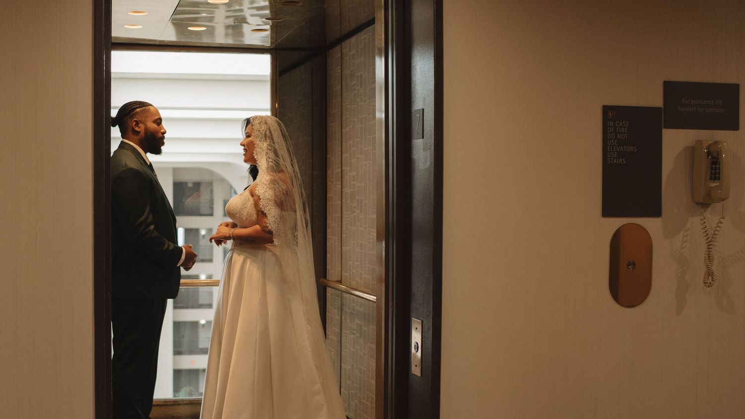 Bride in white gown and groom in black suit stand by elevator doors in elegant wedding venue.
