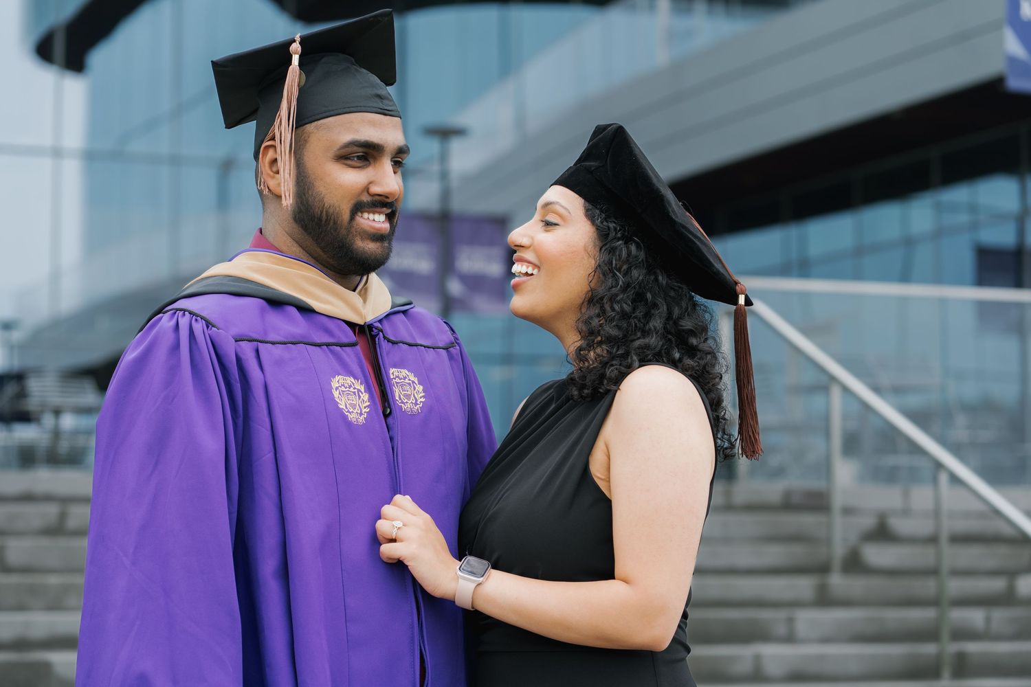 A couple in purple graduation attire and mortarboards smile at each other outside a modern building.