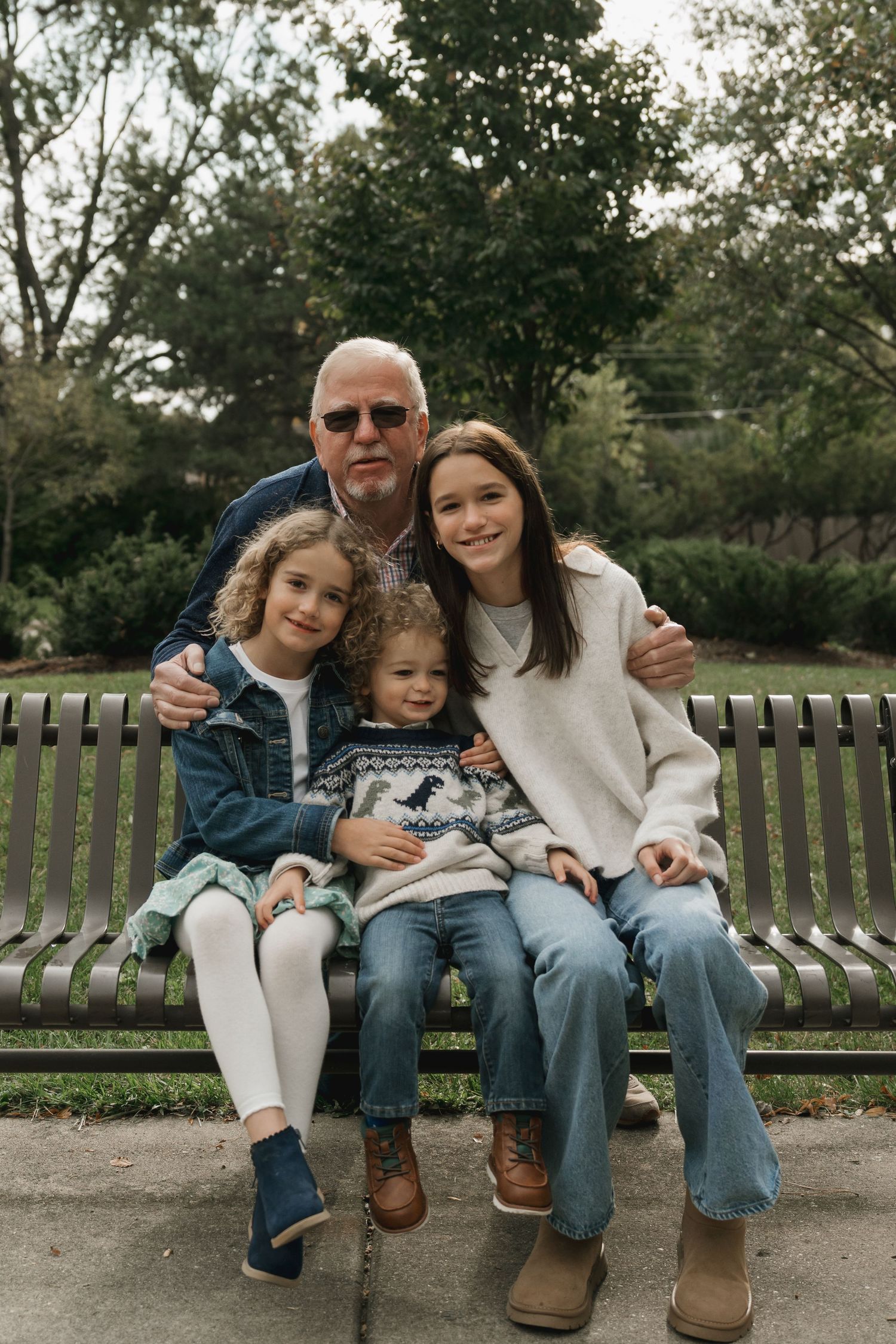 Grandfather sitting on park bench with three grandchildren smiling for portrait.