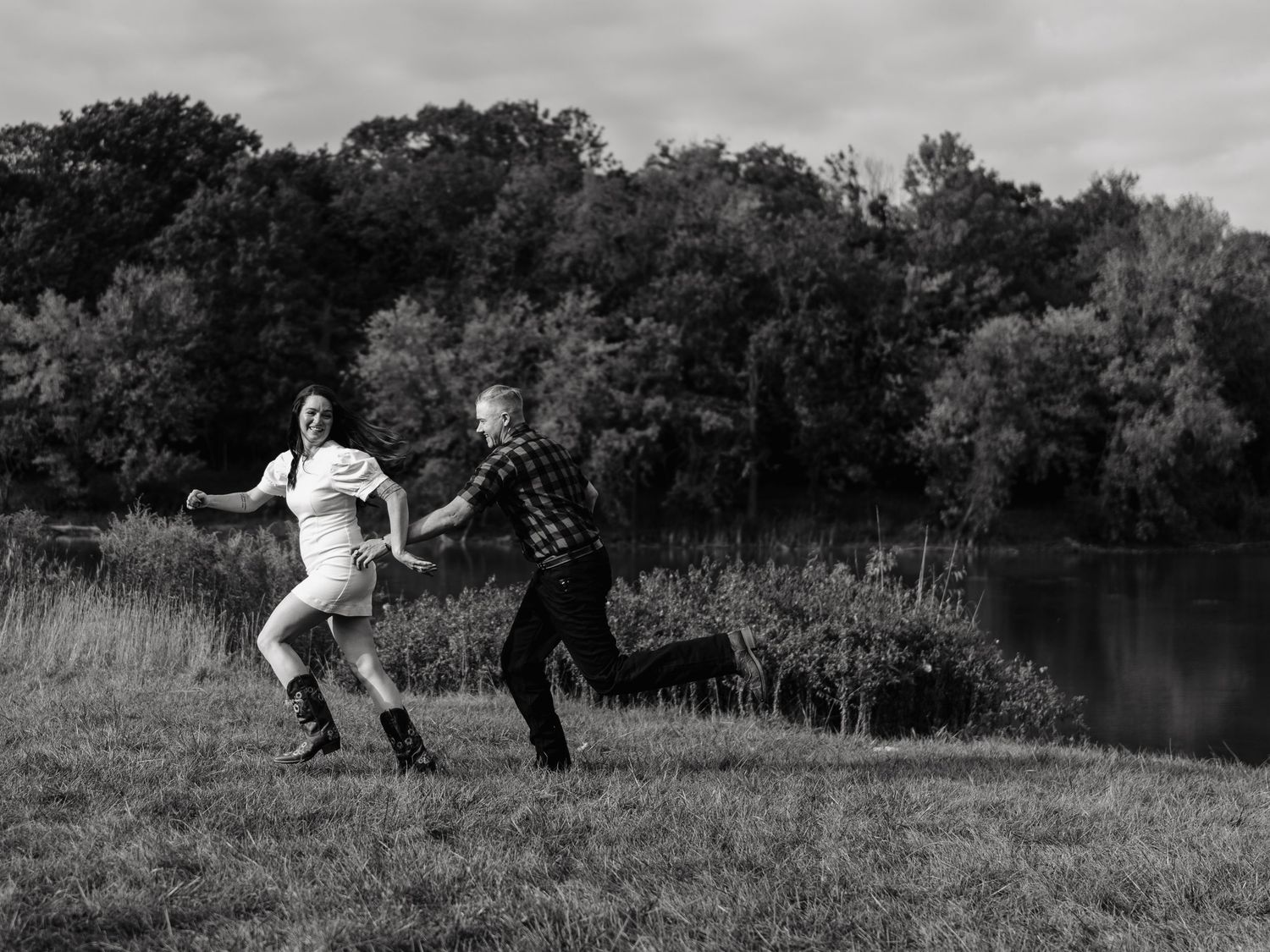 Black and white photo of couple playfully running hand-in-hand along a lakeside meadow.