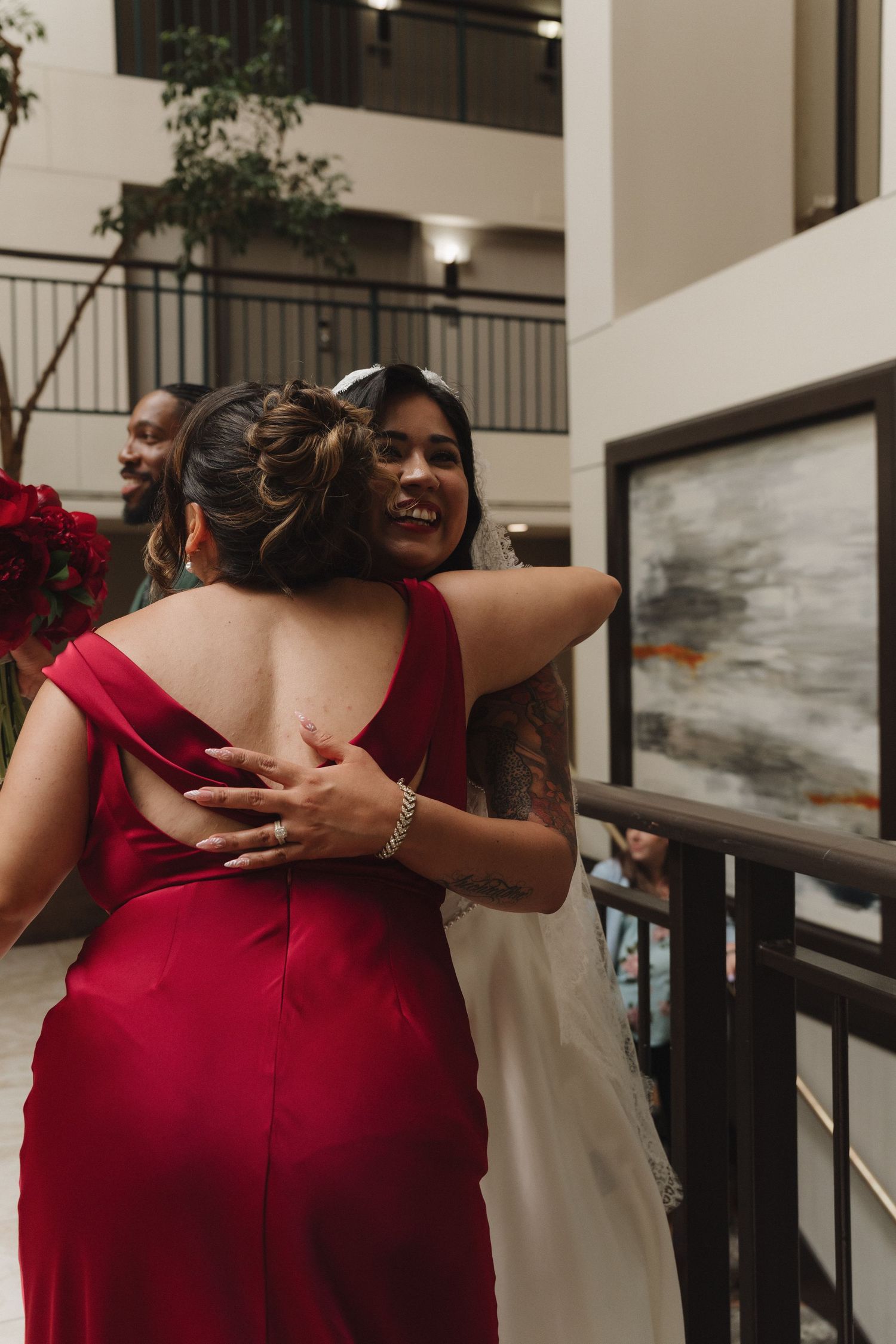 Bride in white gown hugging bridesmaid in red dress at wedding reception venue.