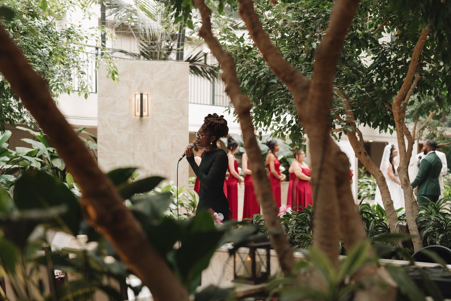 Bridesmaids in red dresses gather among lush green trees before a wedding ceremony.