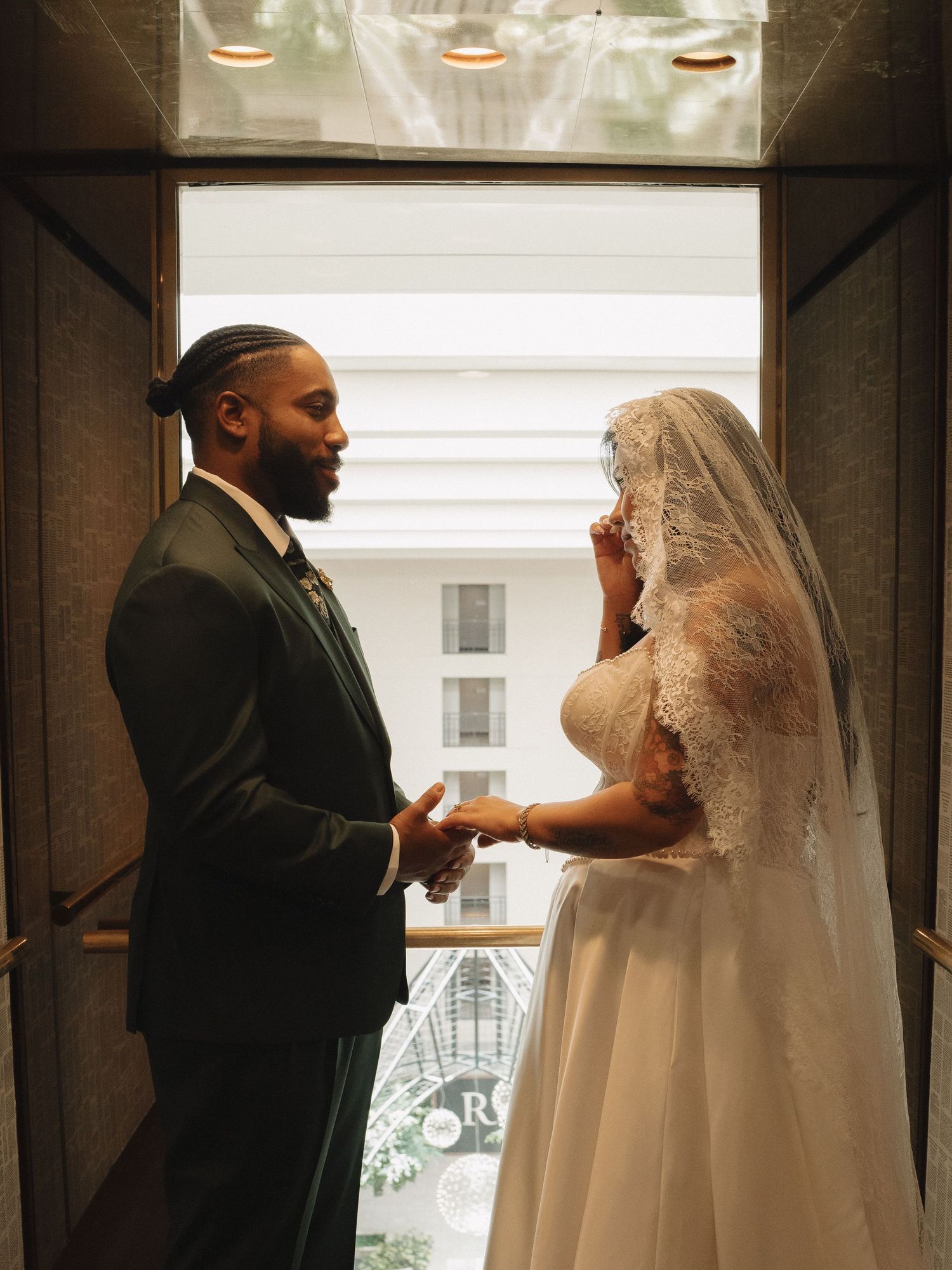 A groom in a dark suit holds hands with a bride in a lace wedding gown and veil, standing in a doorway.