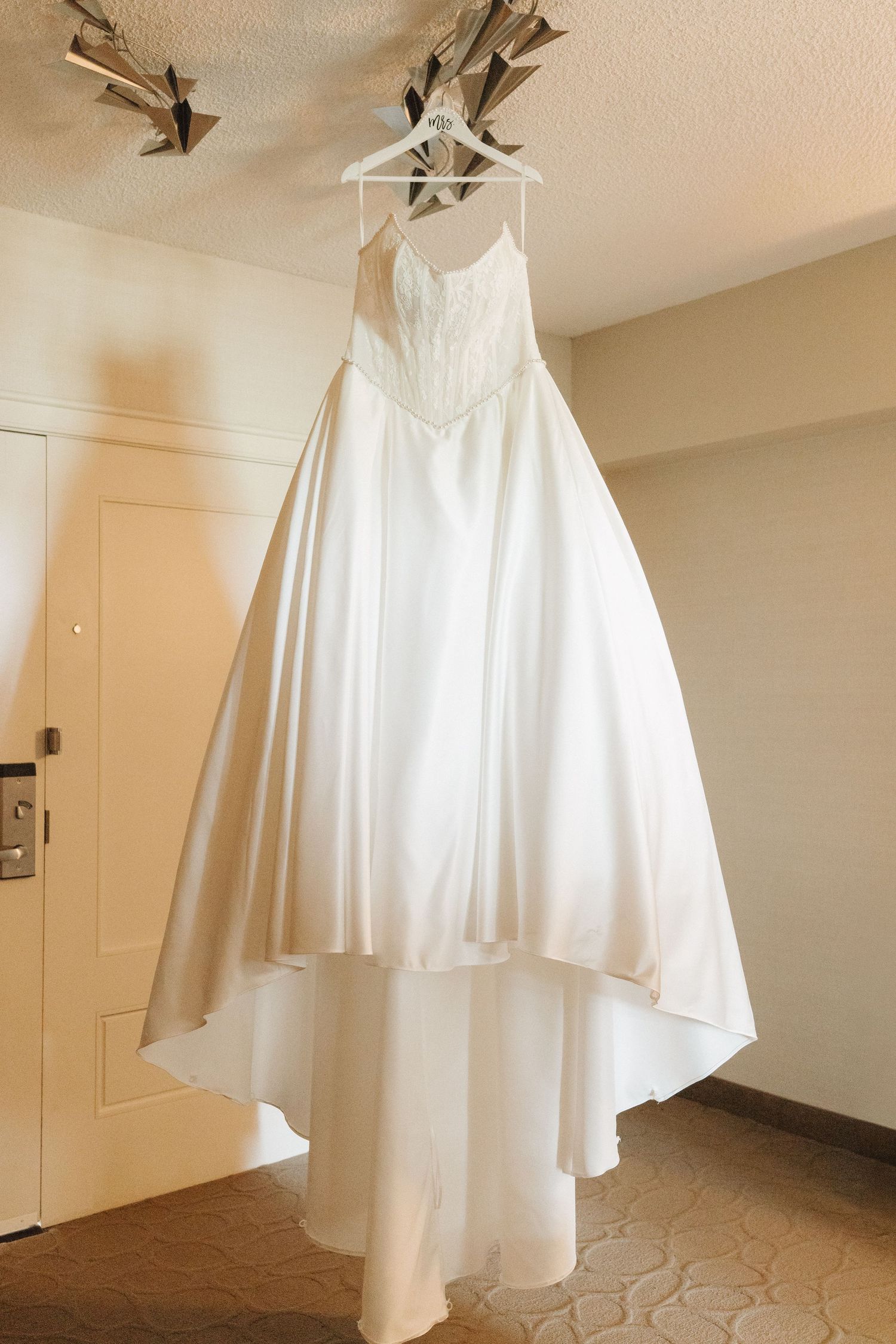 White satin A-line wedding dress with lace bodice hanging on hanger in hotel room before bridal ceremony.