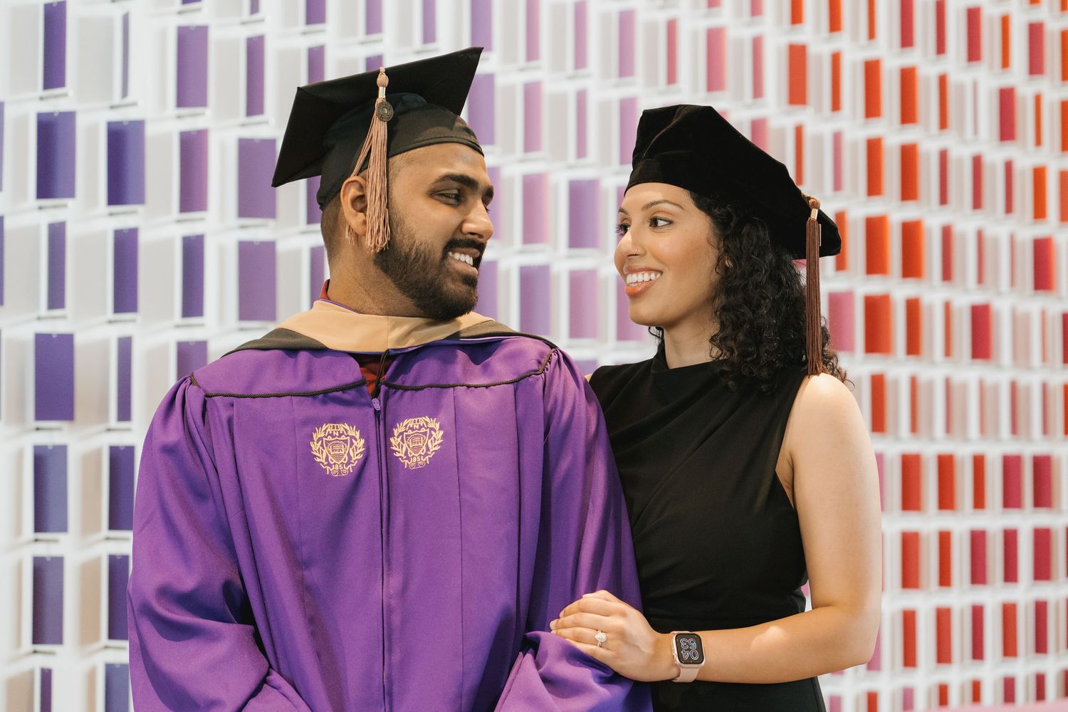 A graduate in purple robes and mortarboard smiles with a woman in black at a colorful backdrop.