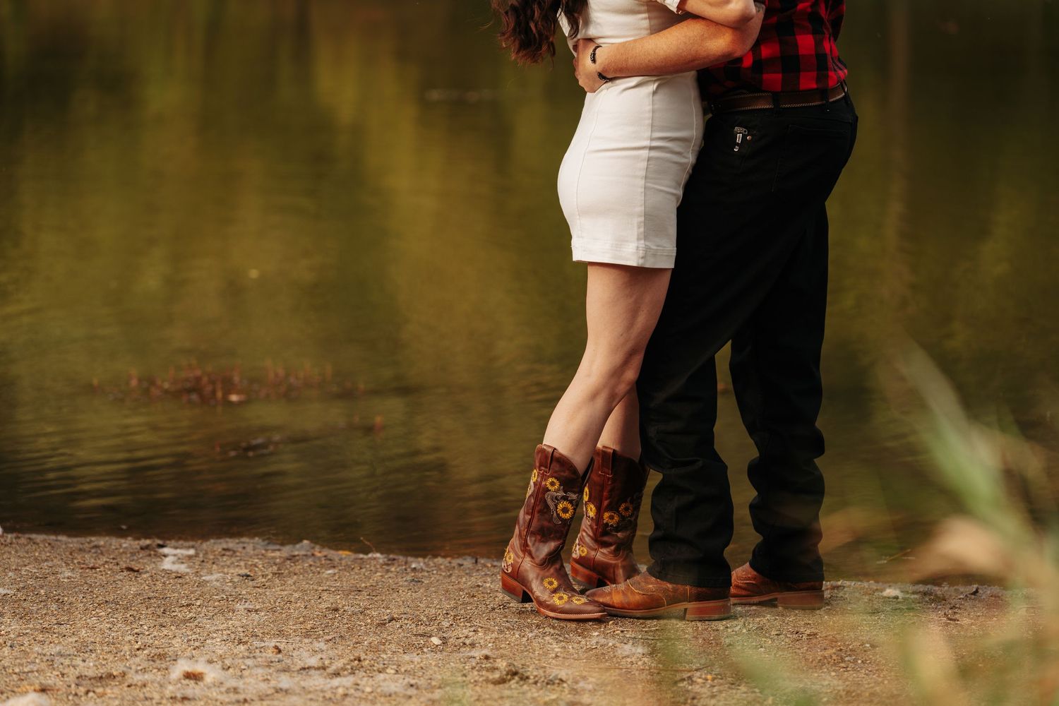 Couple embracing by a lake, woman in white dress and cowboy boots, man in red flannel shirt and black jeans.