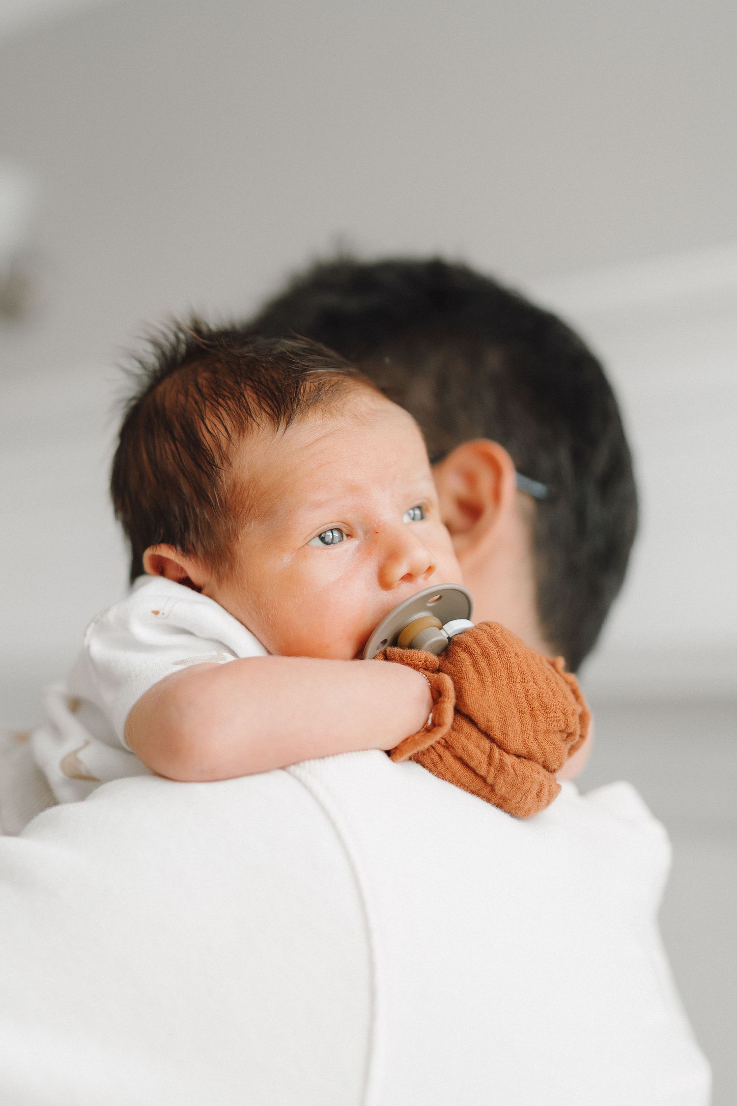 Newborn baby with a pacifier resting on parent's shoulder, holding a rust-colored muslin cloth.