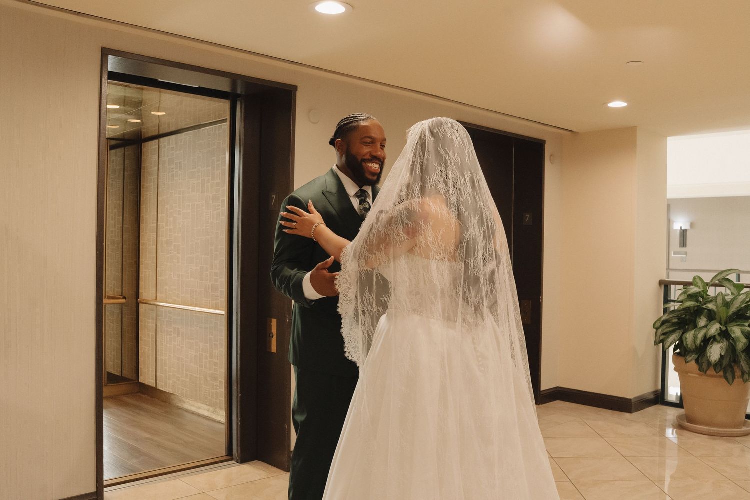 Groom in dark suit reacts with joy seeing bride in white wedding gown and veil during first look moment.