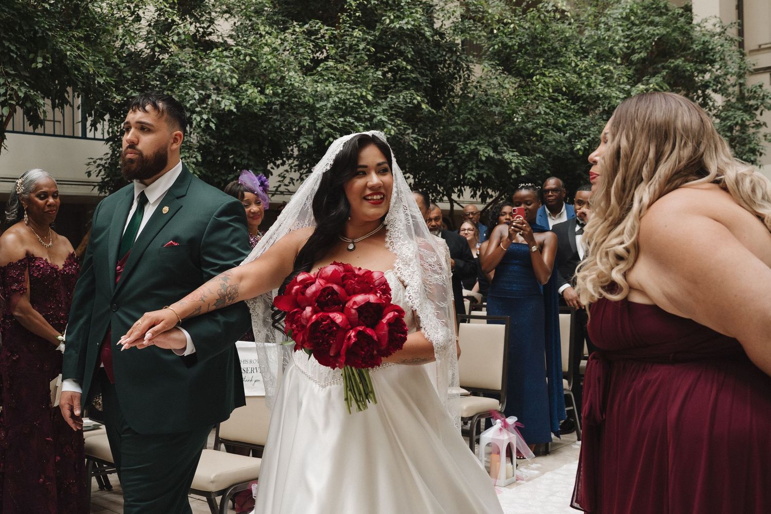 Bride in white gown holding red bouquet walks down aisle flanked by groomsman in green suit and burgundy bridesmaids.