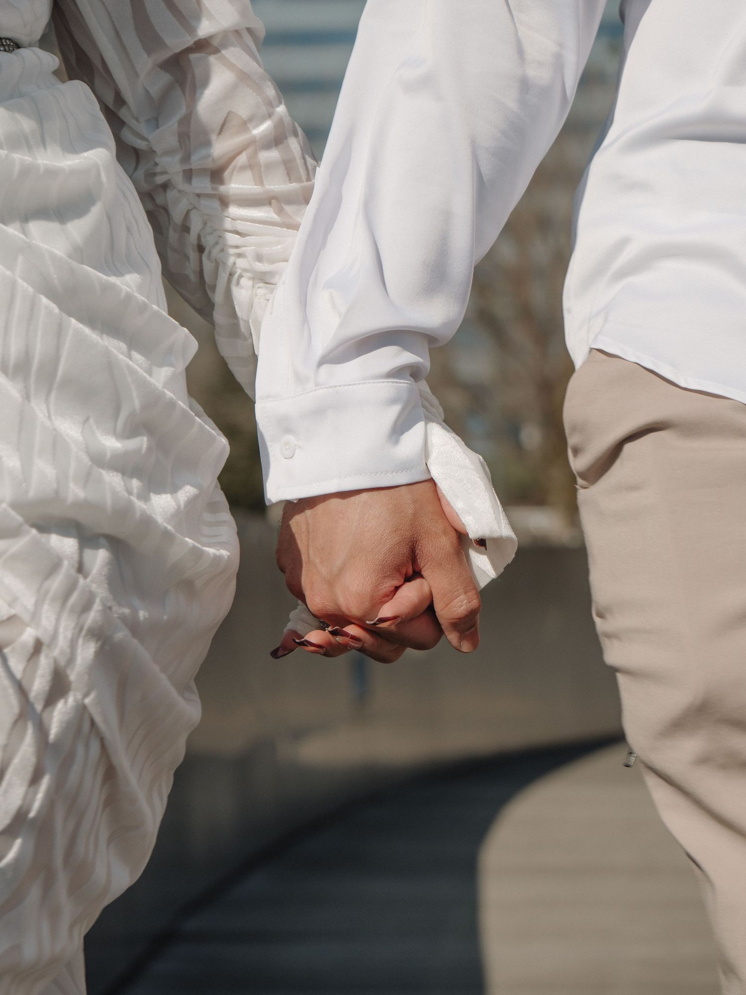 Close-up of a couple holding hands, both dressed in white and beige outfits outdoors.