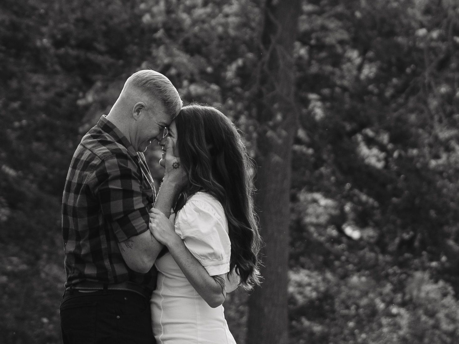 Black and white photo of couple embracing intimately in a wooded forest setting.