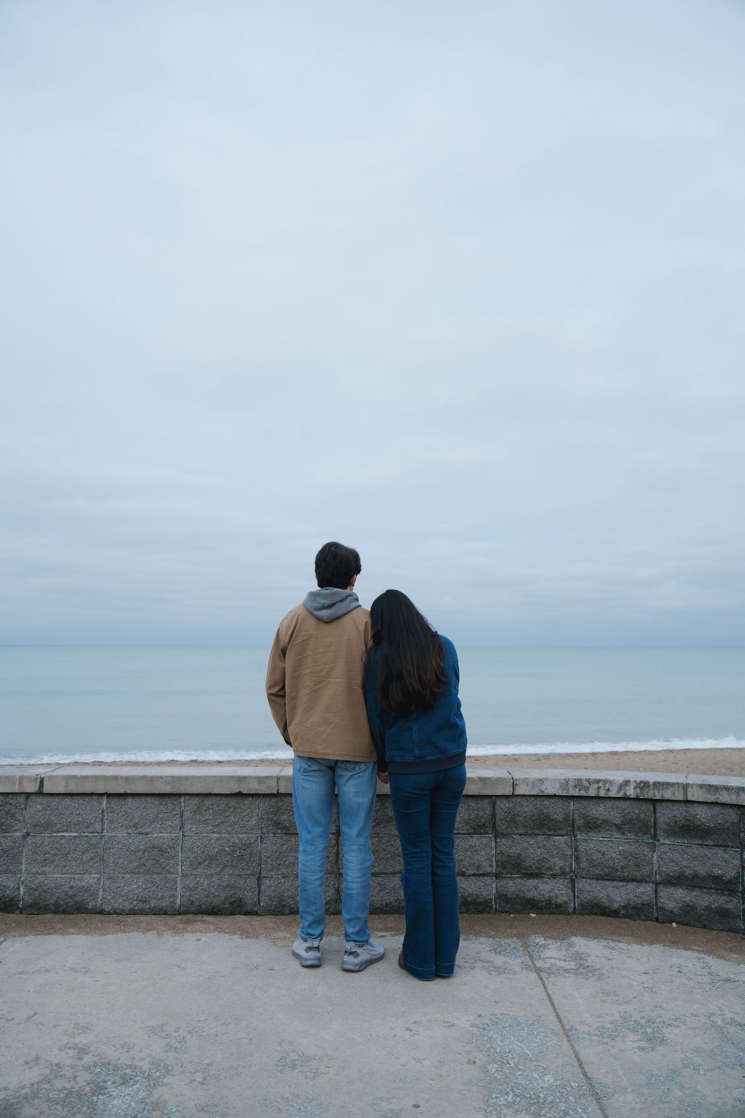 A couple stands together looking out at a calm ocean view on an overcast day.