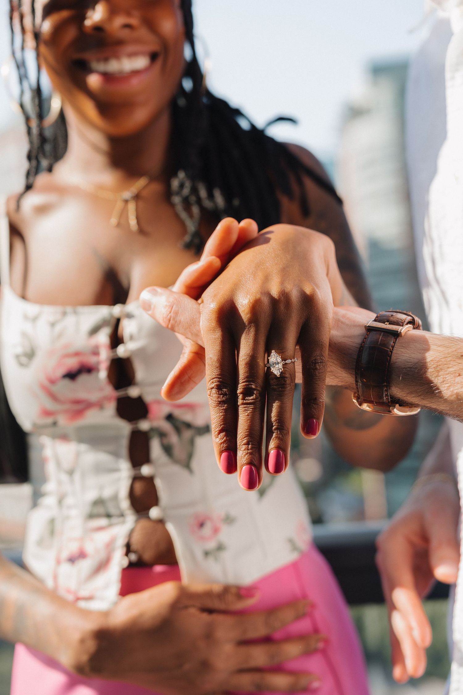 Close-up of woman showing off marquise engagement ring while smiling with city skyline in background.