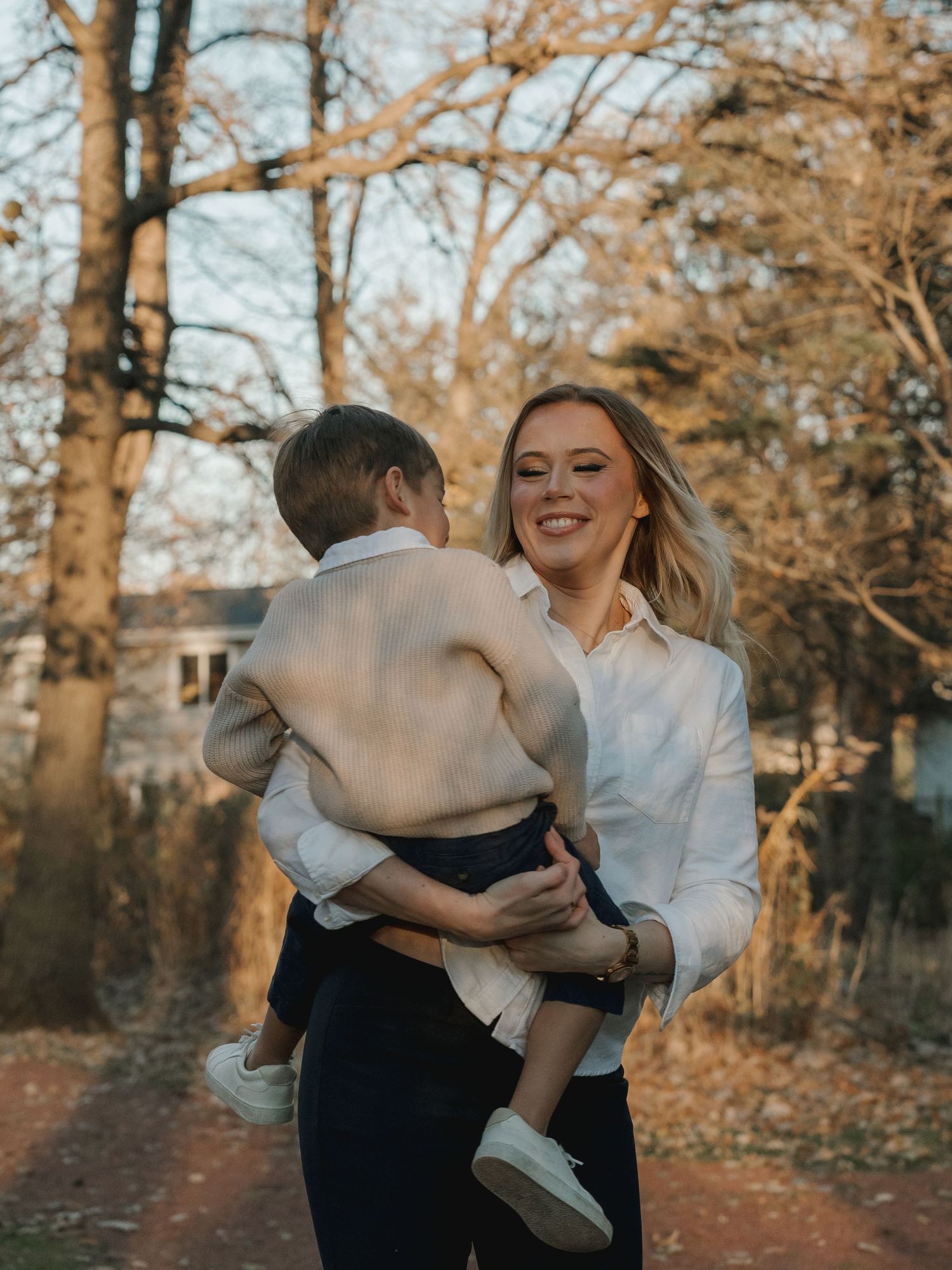 A smiling mom in a white shirt holds her young child outdoors among autumn trees in warm golden light.