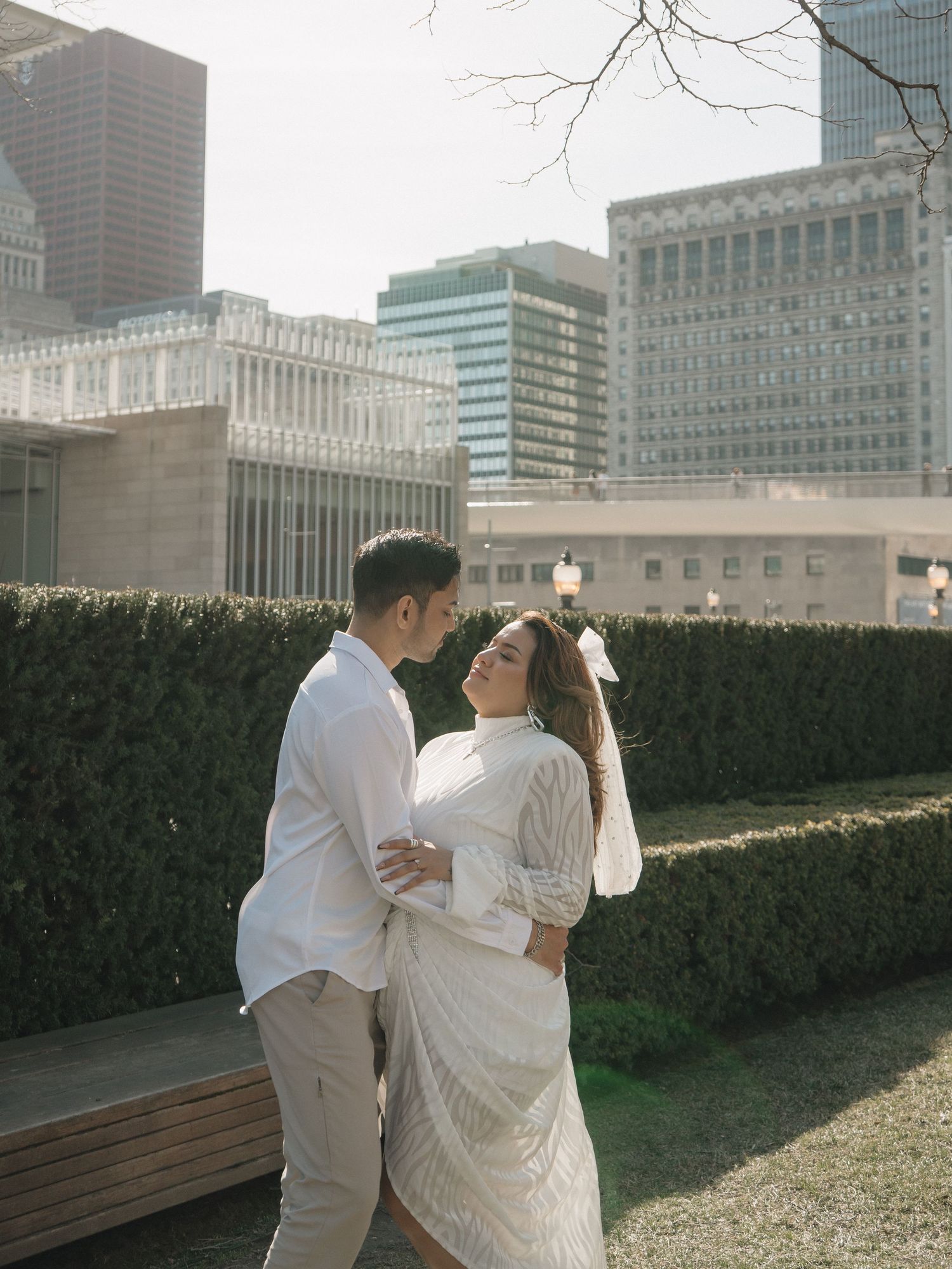 A couple in white outfits share a romantic moment in an urban park with manicured hedges and city skyscrapers behind them.