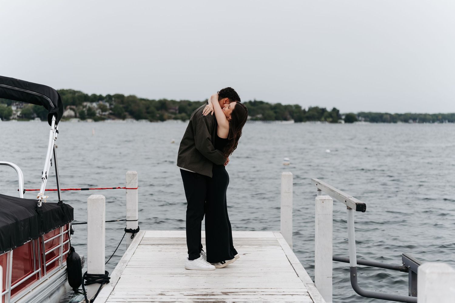 Couple sharing a romantic kiss on a wooden dock by a lake, with a pontoon boat and calm water in the background.