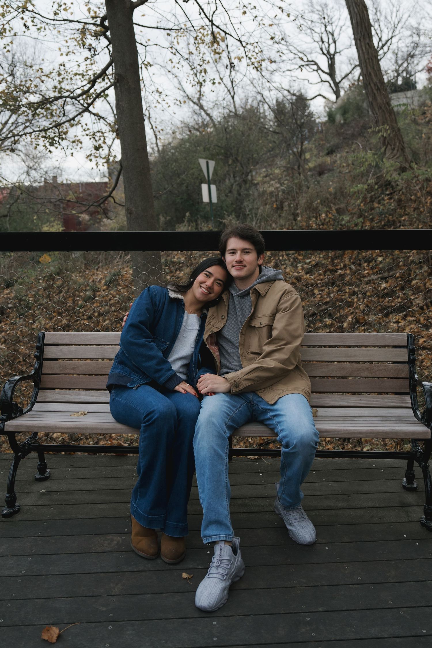 A young couple sits close together on a wooden park bench surrounded by autumn trees.