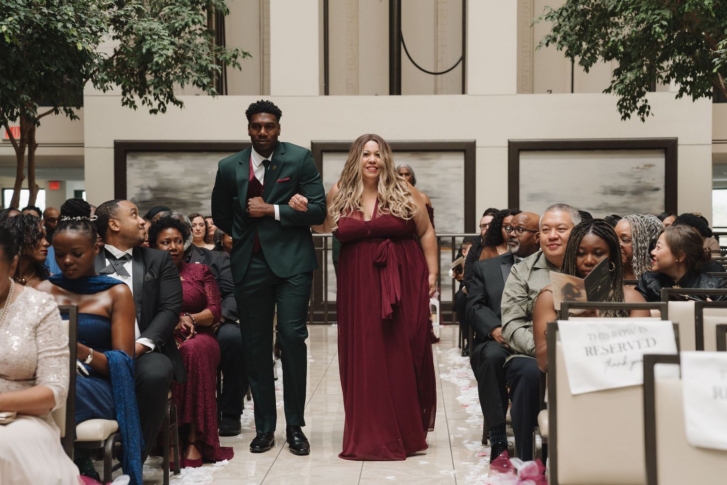 Bridesmaid in burgundy gown walks down the aisle with groomsman in dark suit at indoor wedding.