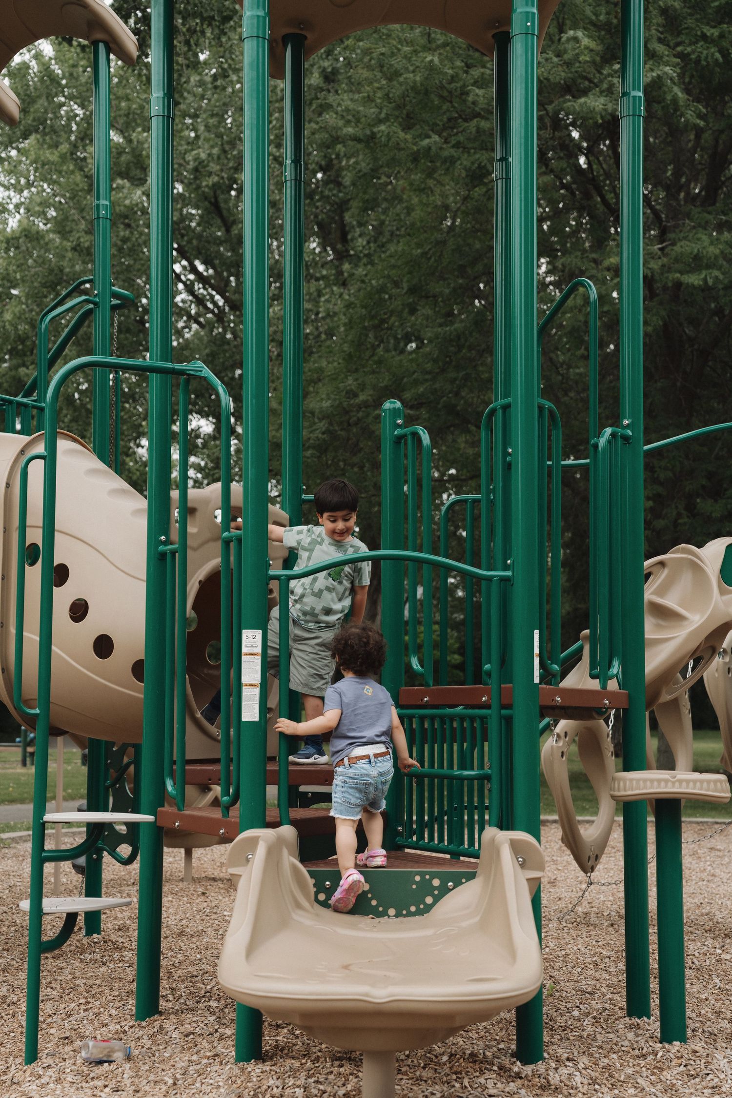 Two children playing on a green and tan playground structure in a park surrounded by trees.