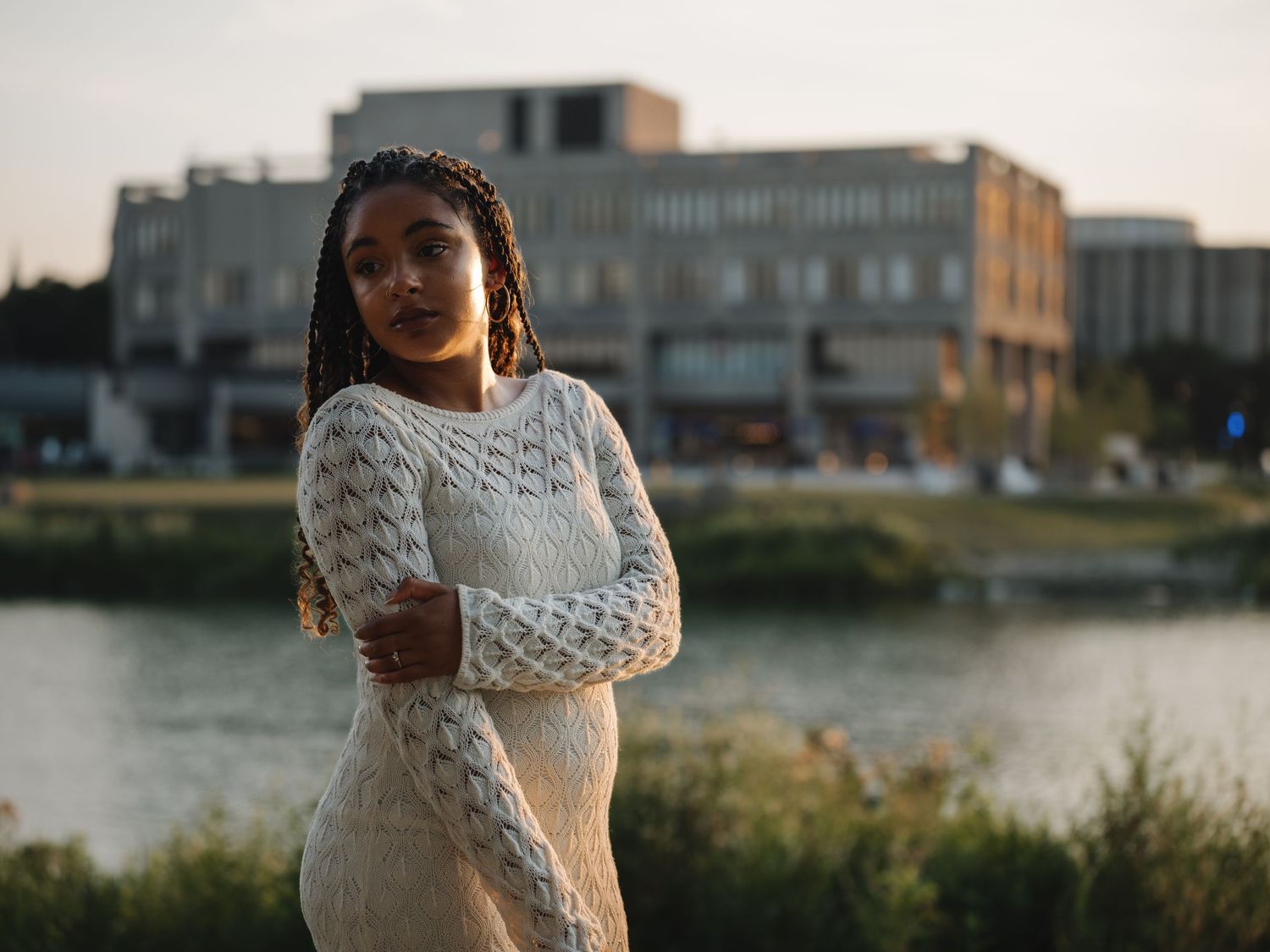 Young woman in white lace dress standing by a river with urban buildings in the golden hour background.