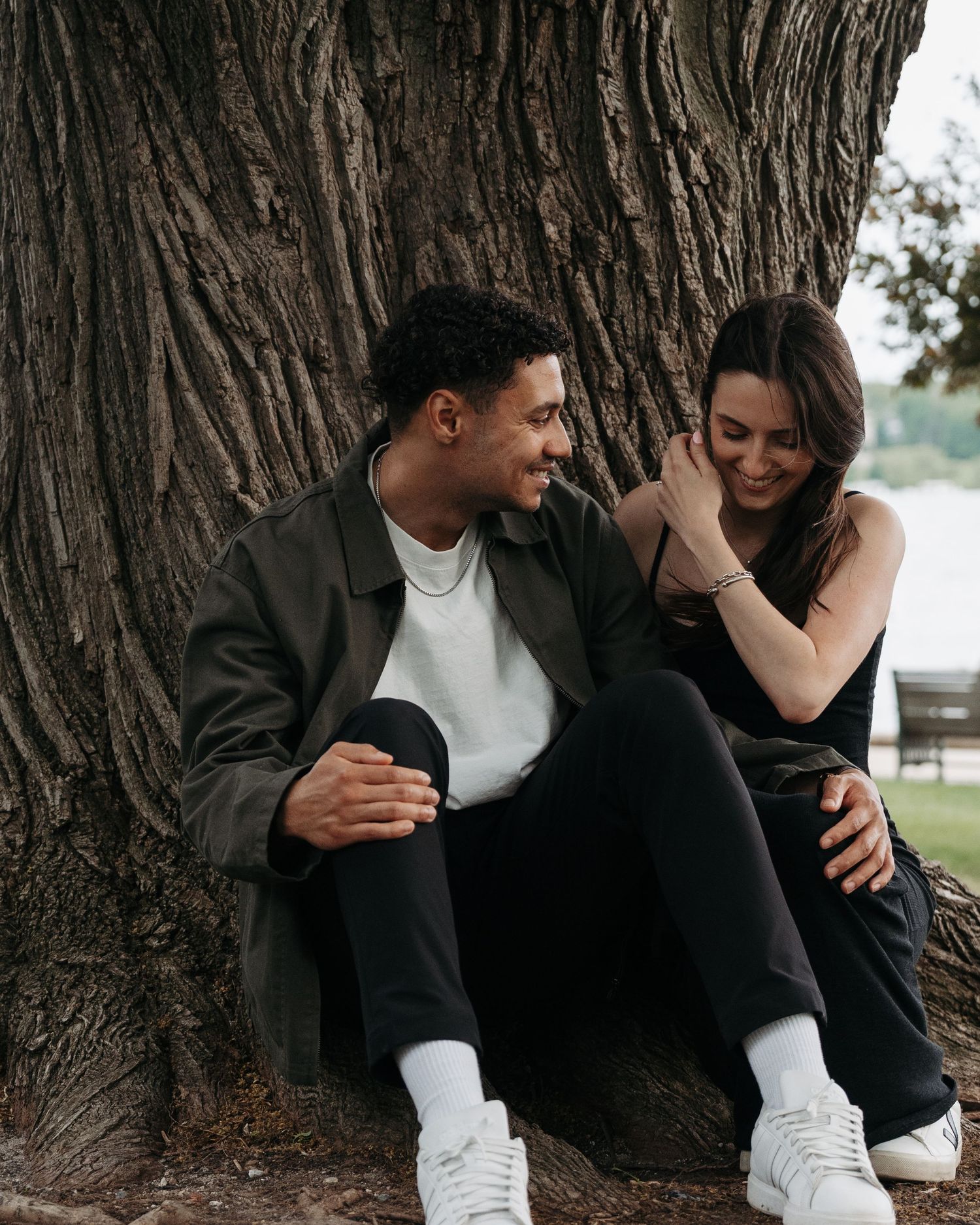 A young couple sitting together against a large tree trunk, smiling and sharing a joyful moment outdoors.