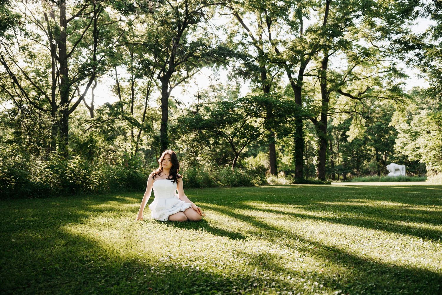 Young woman in white dress sitting cross-legged on grass in a sunlit green park.