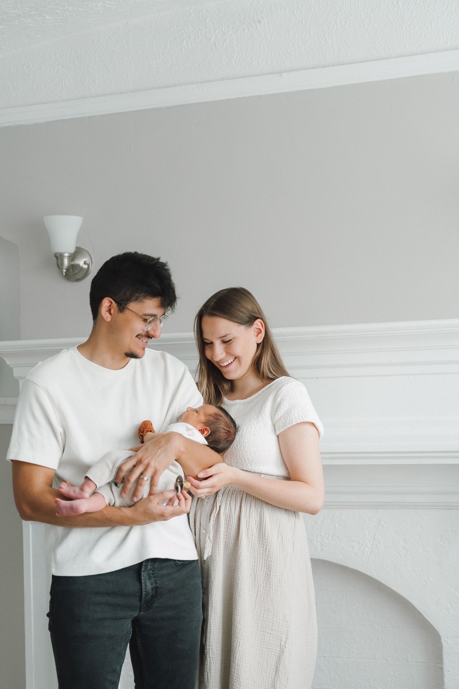 Happy new parents smiling and holding their newborn baby in a bright white living room.