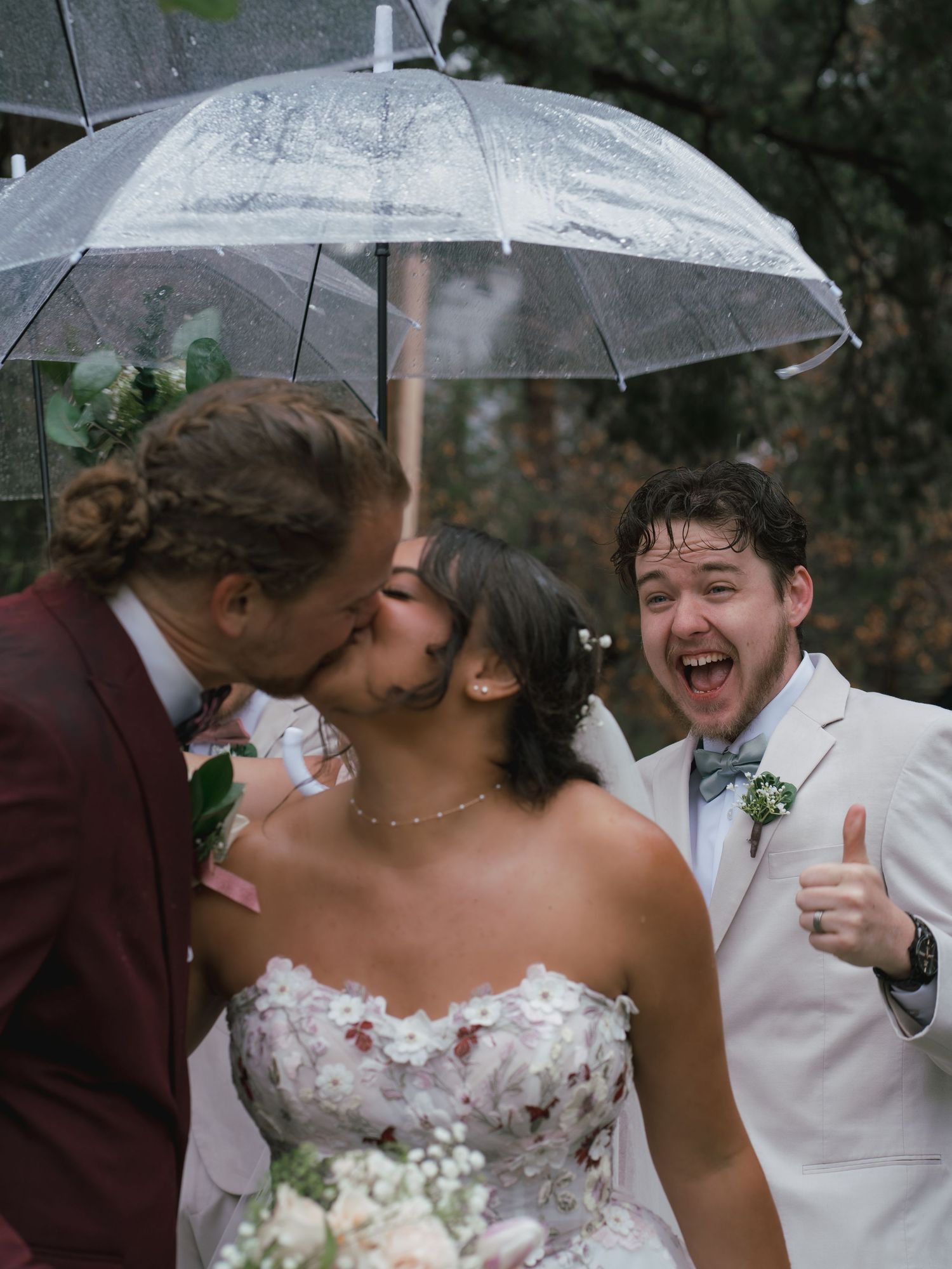 Groom kisses bride under umbrella as excited groomsman gives thumbs up at rainy outdoor wedding.