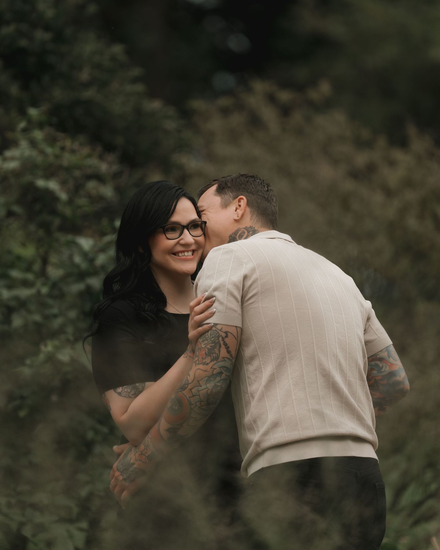 A tattooed couple shares a tender kiss outdoors, surrounded by lush green foliage, as the woman smiles warmly.