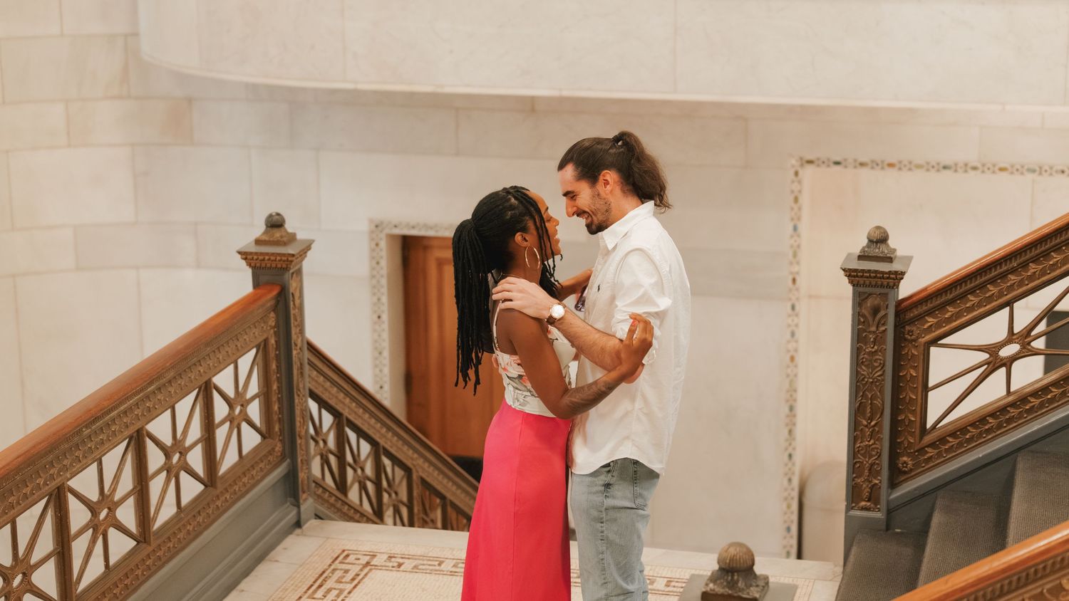 A couple embraces romantically on an elegant staircase, the woman in a red skirt and the man in a white shirt.