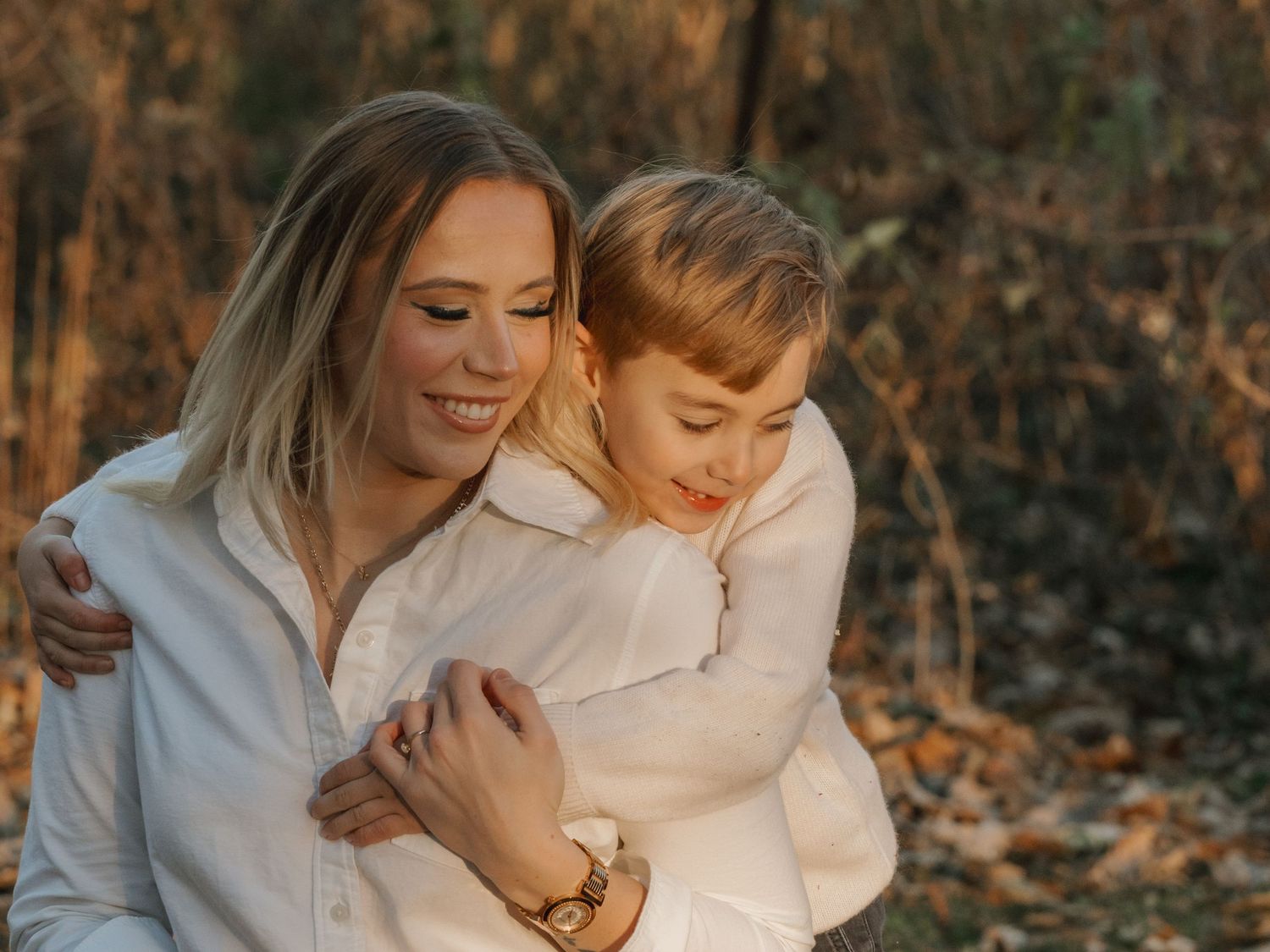 Smiling blonde mother hugs her young son in matching white shirts in autumn park.