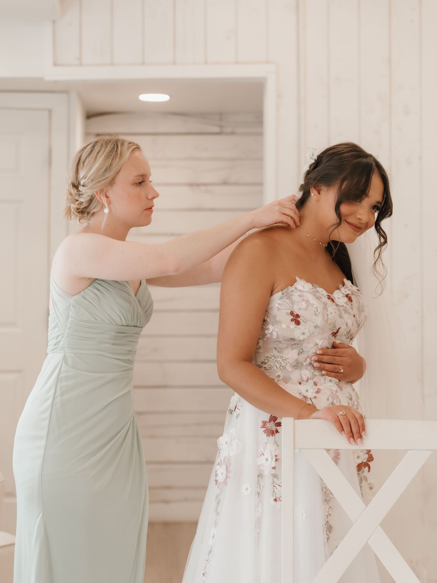 Maid of honor in sage green dress helps bride in floral wedding gown with hair accessories before ceremony.