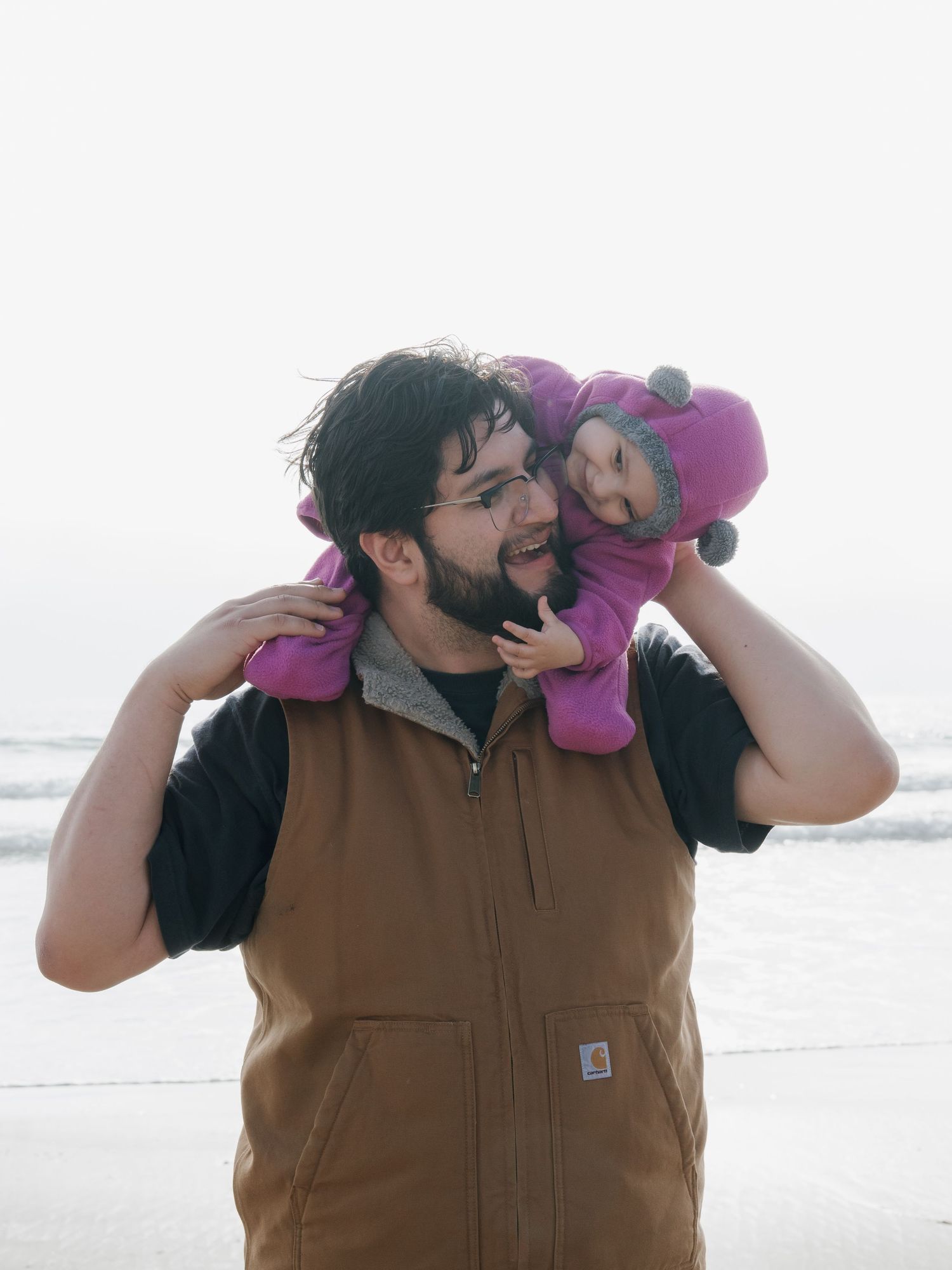 A bearded man in a Carhartt vest holds a baby in a purple outfit on his shoulders at a foggy beach.