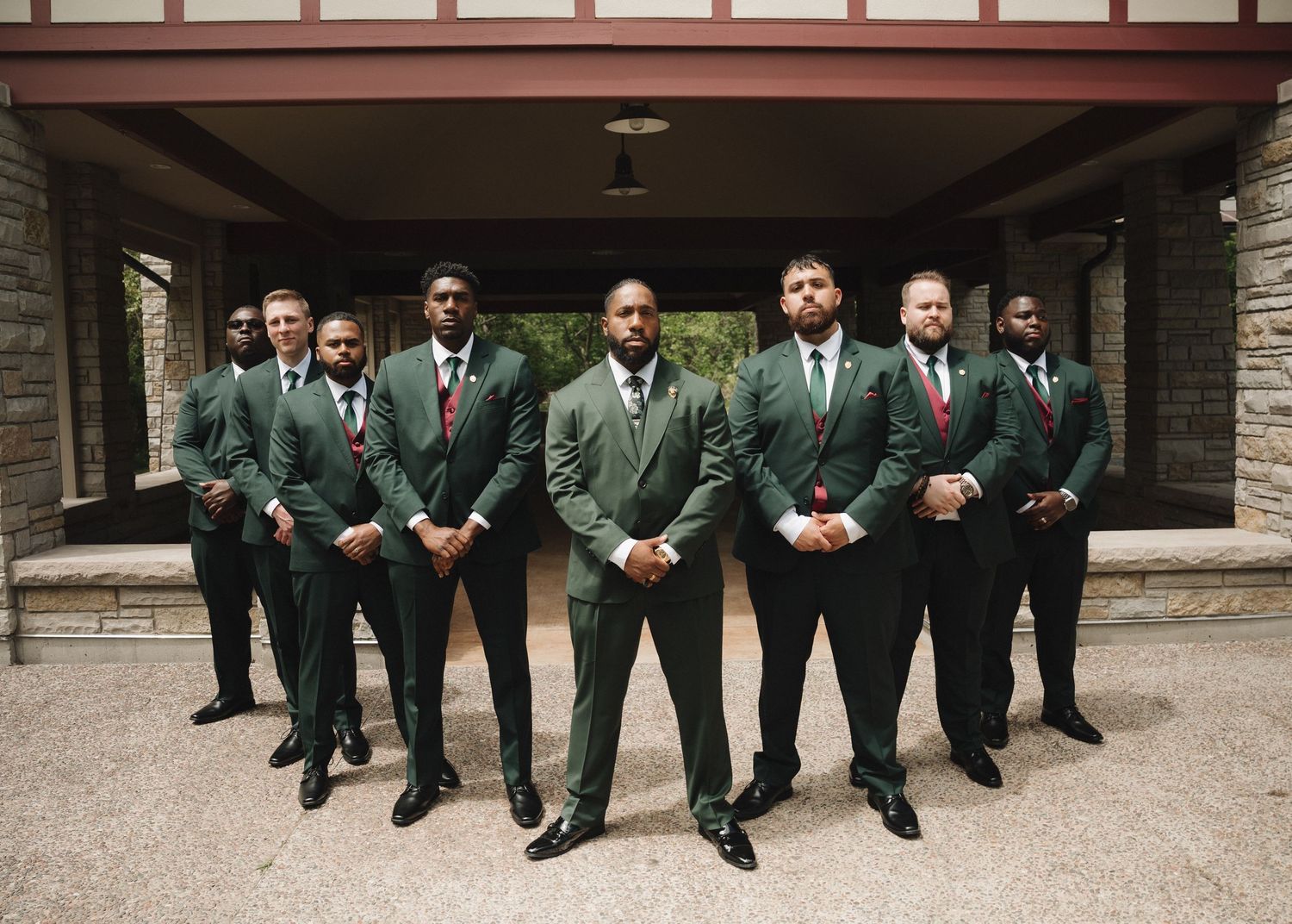 Seven groomsmen in matching dark green suits with red ties pose formally outside a stone building.