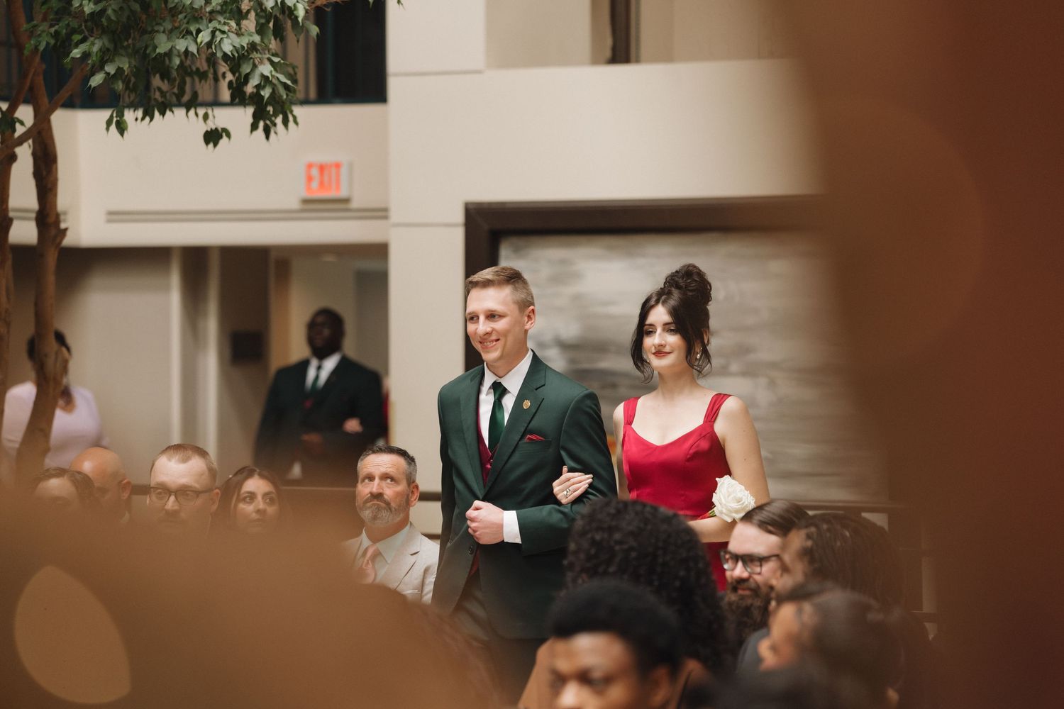 Elegant couple in formal attire, man in green suit and woman in red dress, walking through a wedding venue.