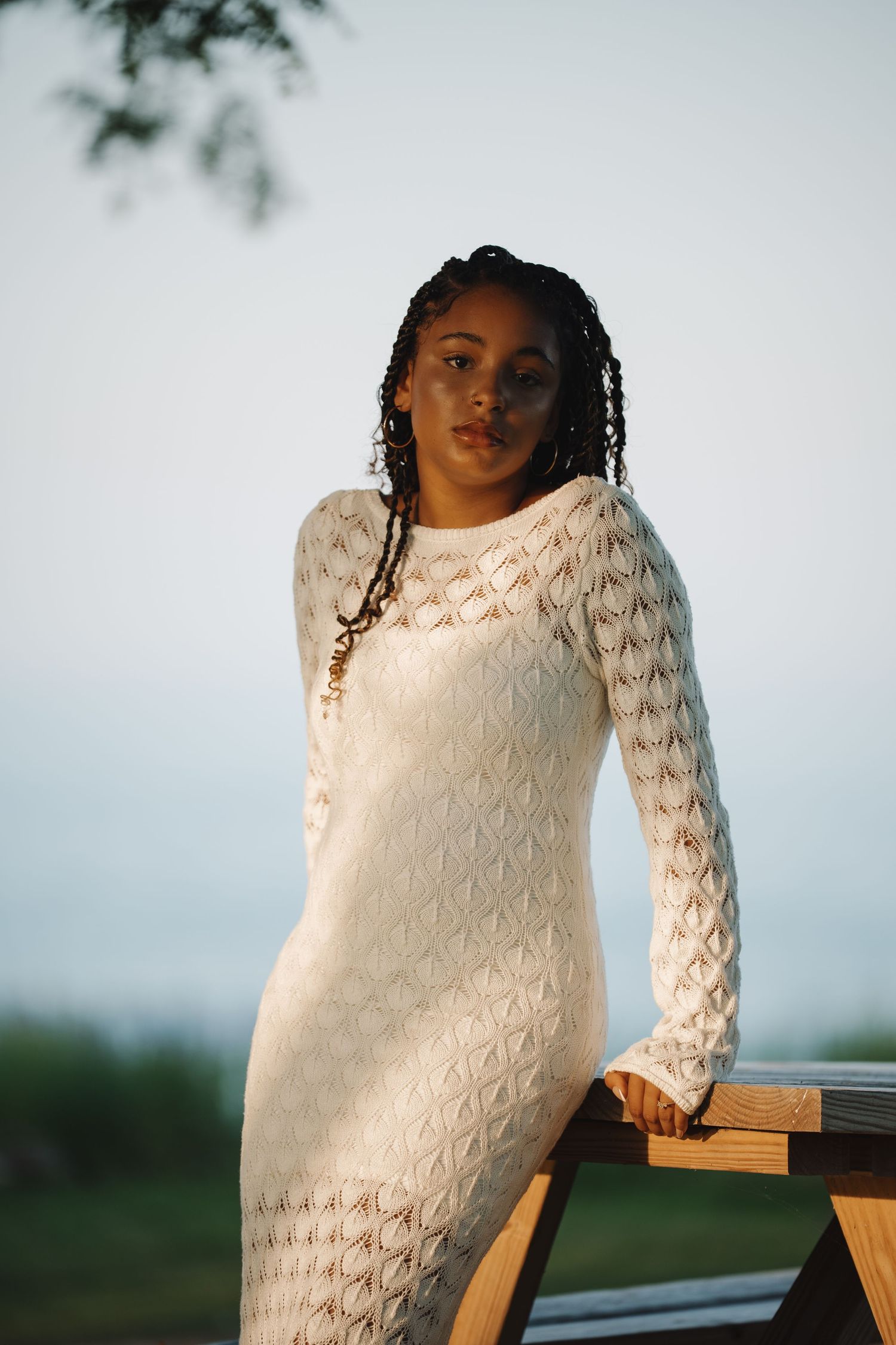 Young Black woman in white lace long-sleeve dress poses outdoors at golden hour, braided hair, leaning on wooden railing.