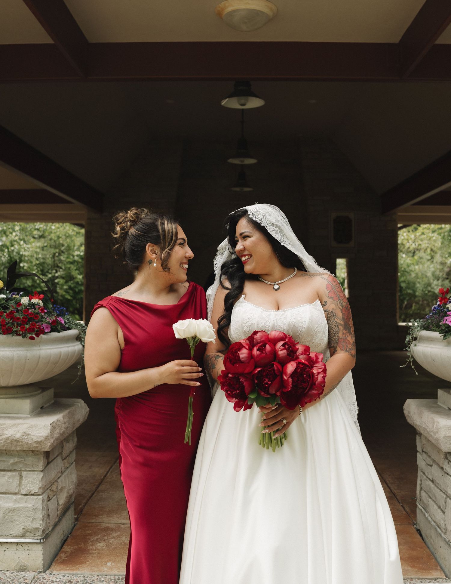 Bride in white gown with red bouquet stands beside woman in red dress, both smiling at each other under a pavilion.