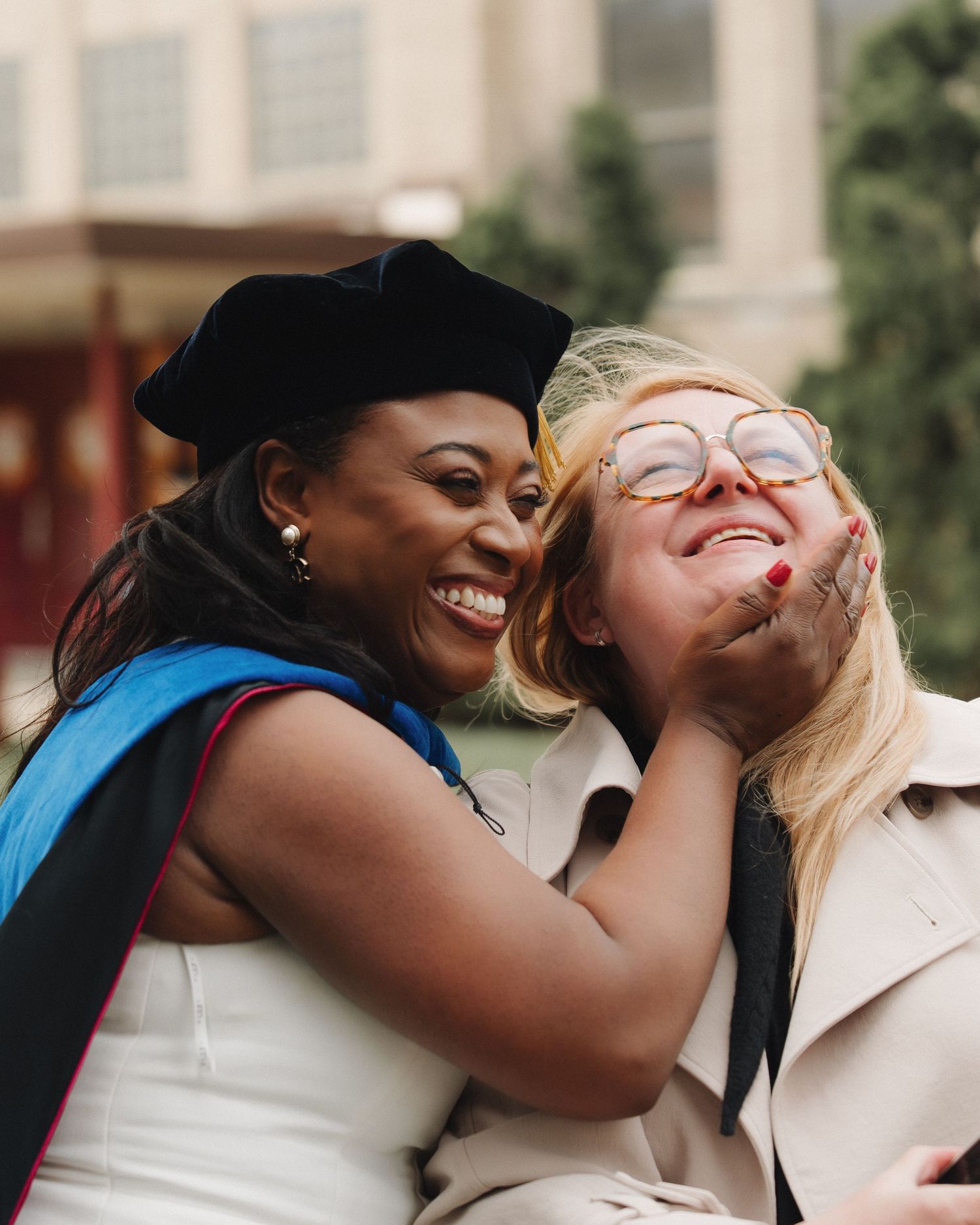 A graduate in cap and gown joyfully embraces a smiling woman in glasses at a graduation ceremony.