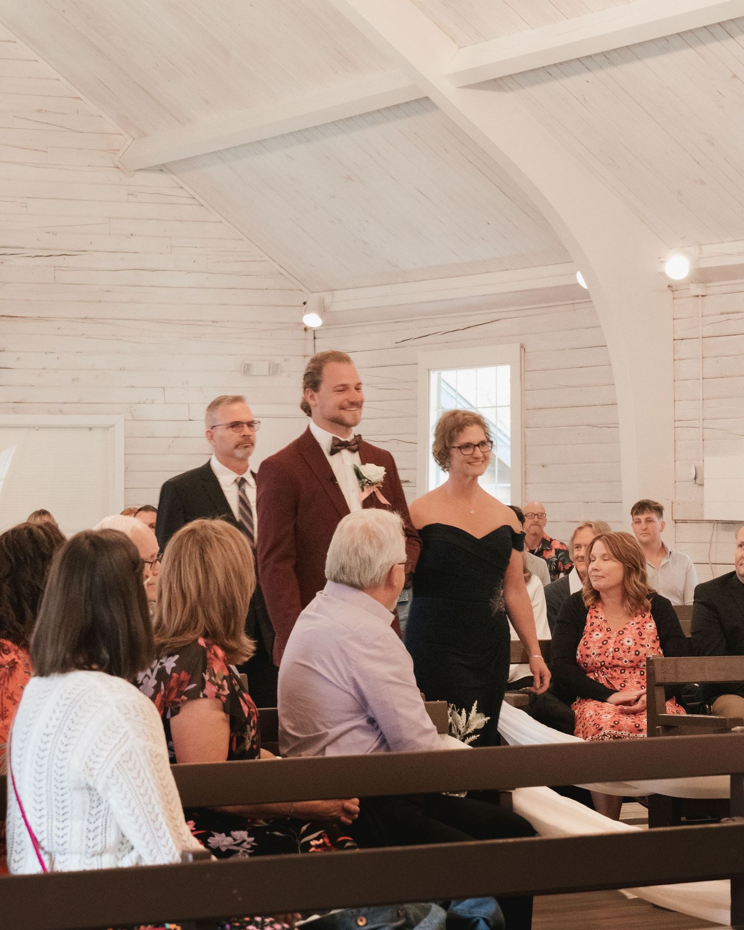 Groom in burgundy suit walks down aisle with mother in black dress at chapel wedding.