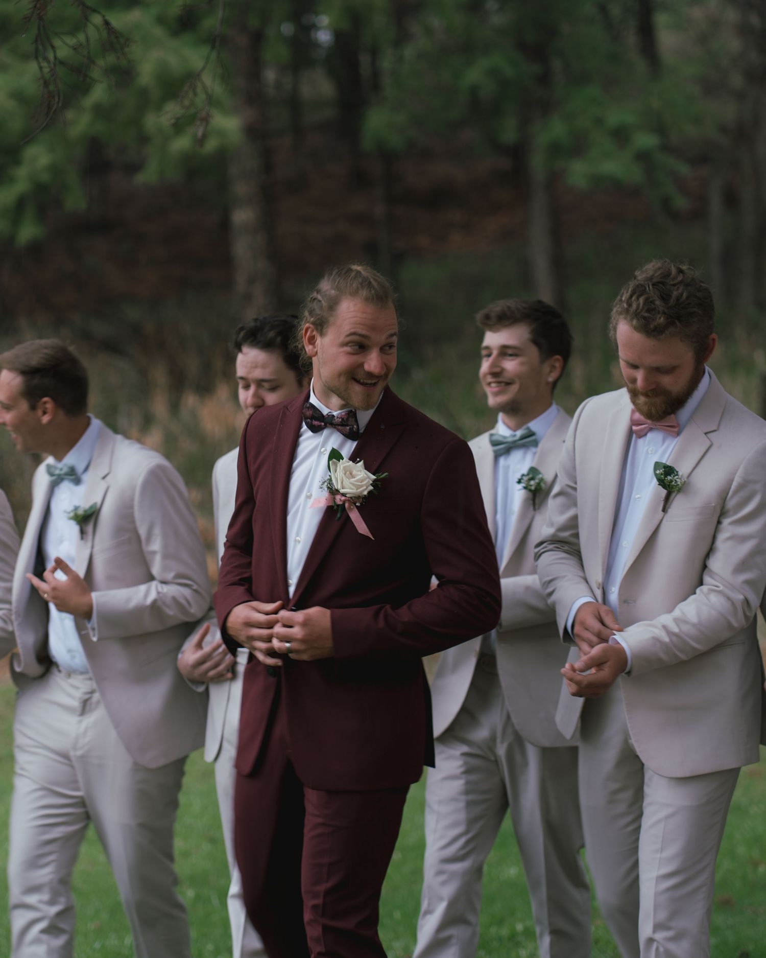 Groom in burgundy suit laughs with groomsmen dressed in light grey suits outdoors.