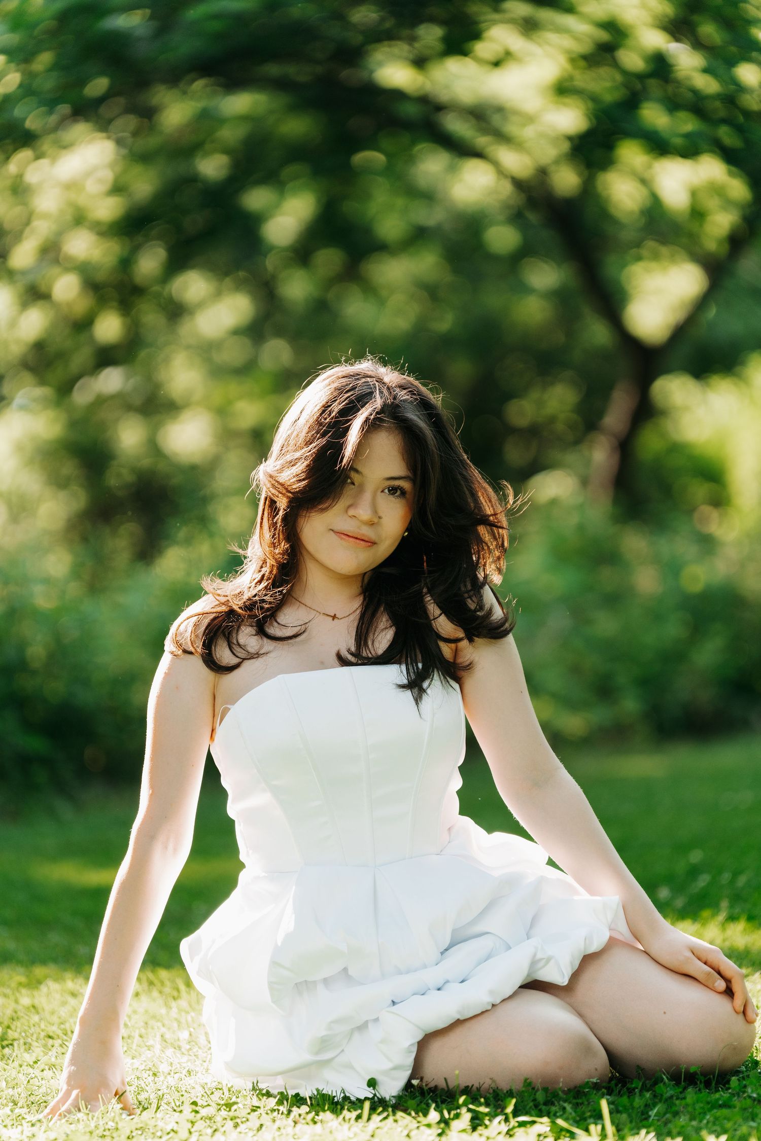 Young woman in white strapless dress sitting cross-legged on green grass in sunny park.