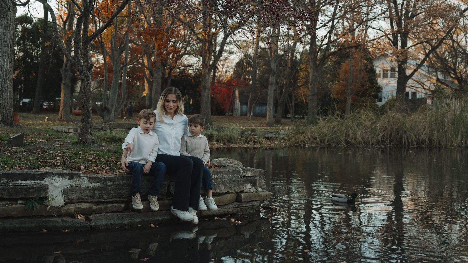 Mother sits with two young children on stone ledge beside a calm autumn pond.