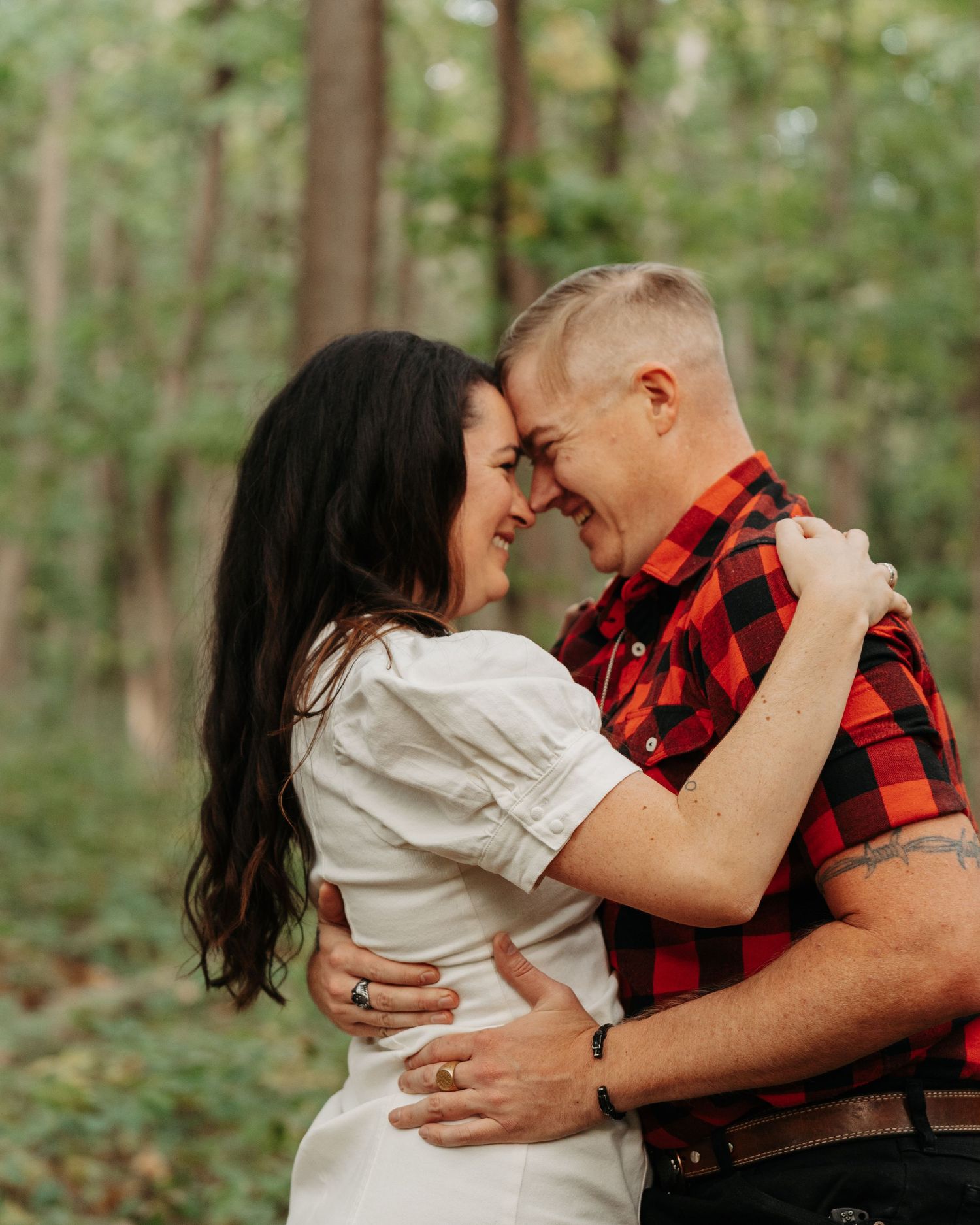 A couple embraces and smiles forehead-to-forehead in a lush green forest, wearing a white dress and red plaid shirt.